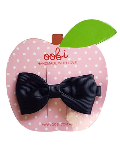 A child's Essentials black Large Bow hair clip attached to a pink polka-dotted apple-shaped card labeled "oobi handmade with love.