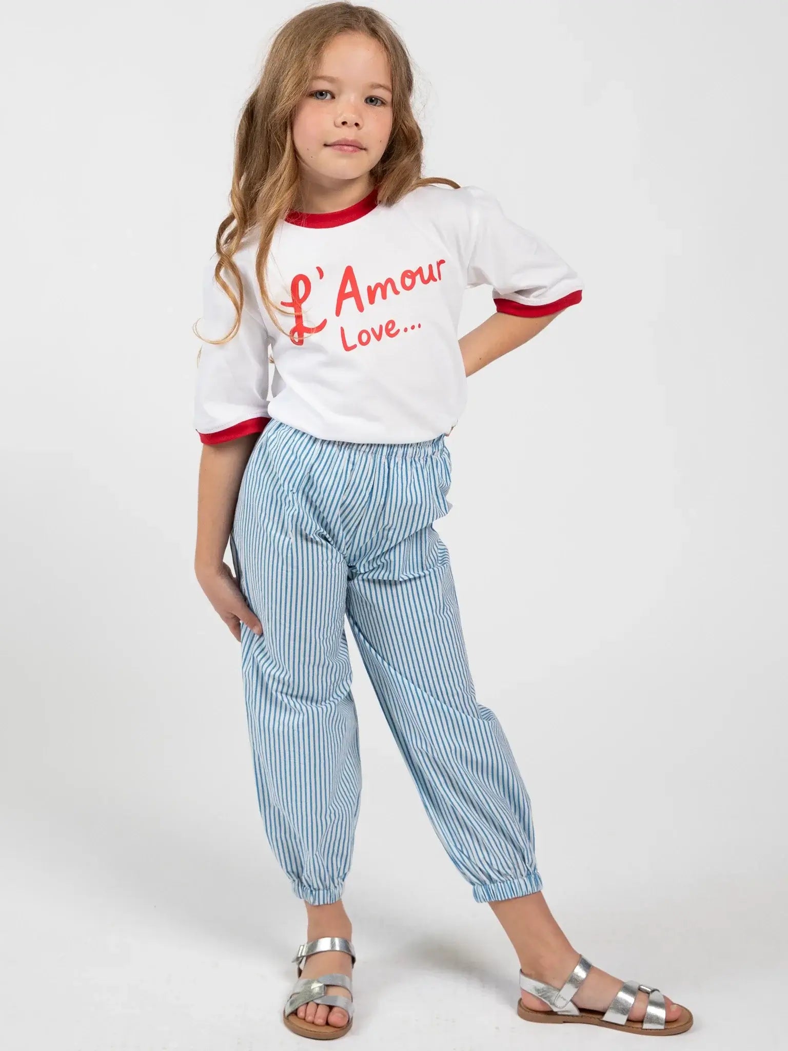 A young girl stands against a plain background, wearing a white "L'Amour Love..." girls tee, Blue Stripe Balloon Pants by Blue Stripe, and silver sandals.