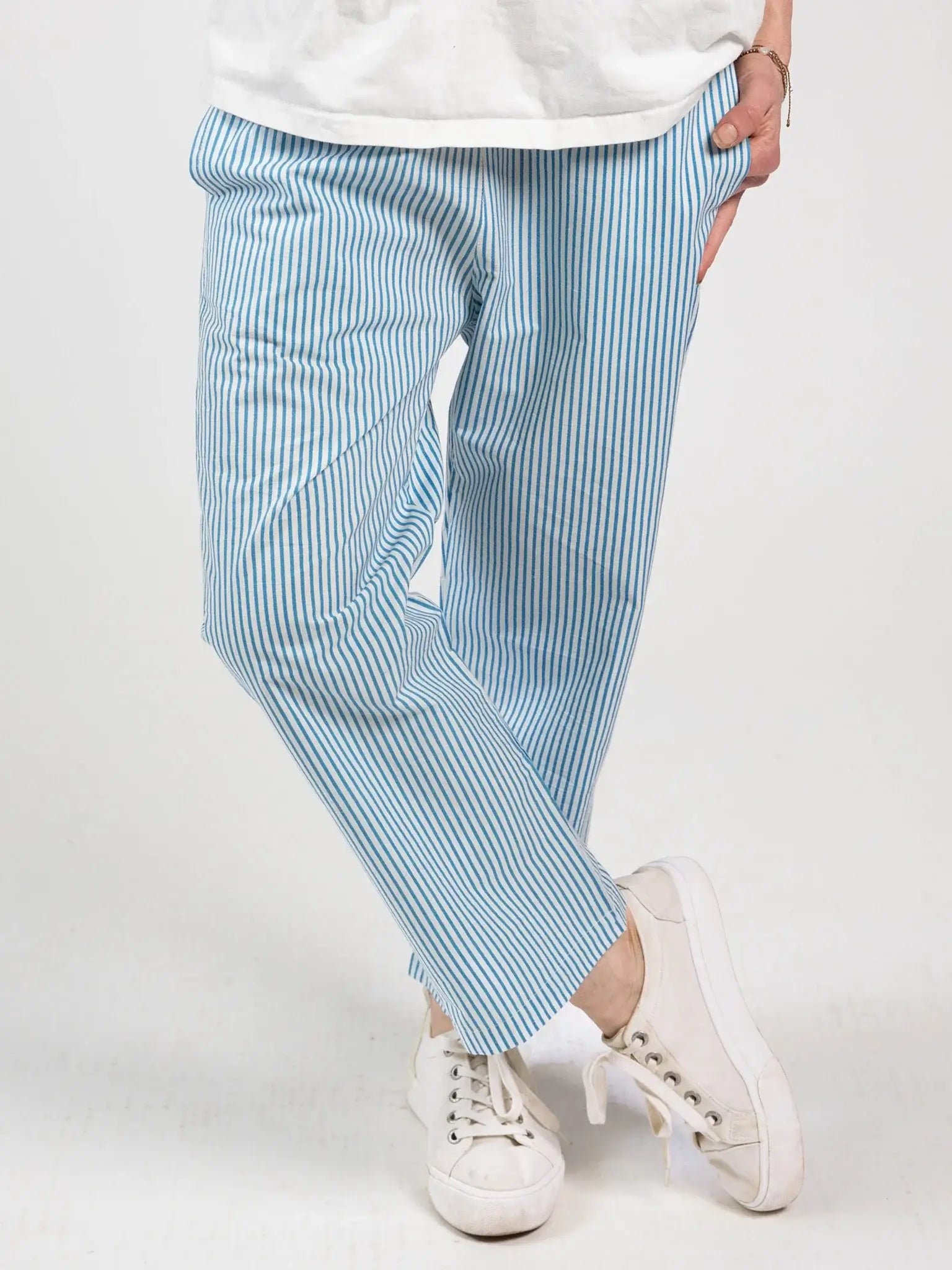 A person wears Blue Stripe Women's Everyday Pants by Blue Stripe—light blue and white vertically striped, with an elastic waistband—paired with a white shirt and white sneakers, standing cross-legged against a plain background.