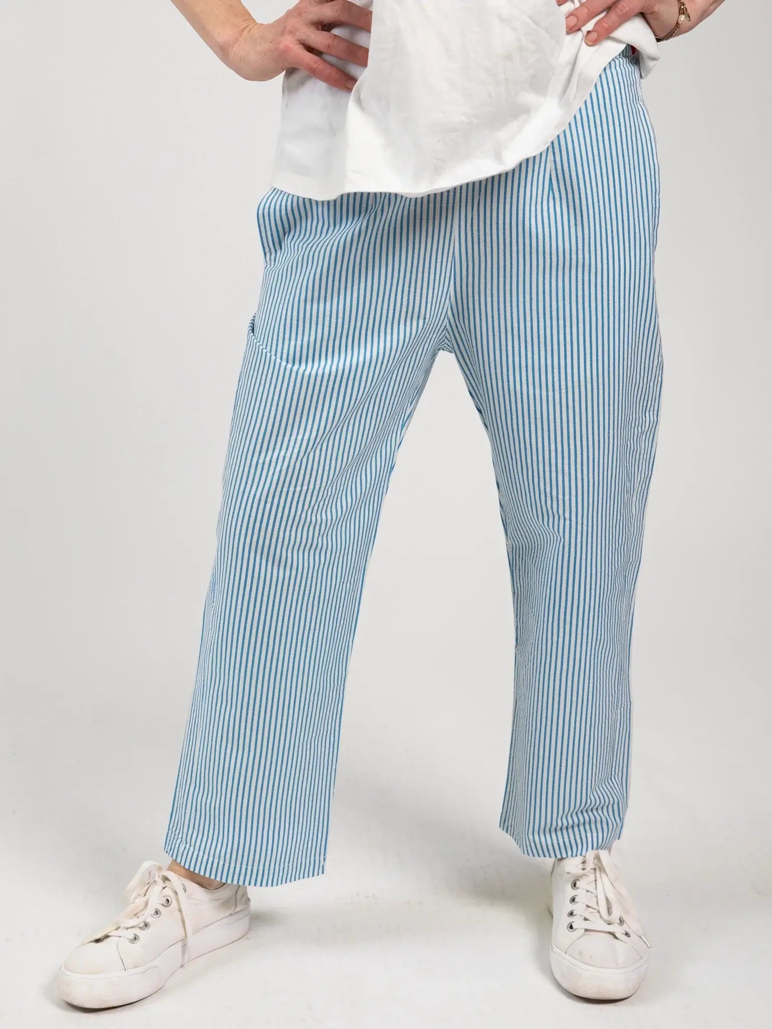 A model wears Blue Stripe Women's Everyday Pants by Blue Stripe—blue and white vertically striped cotton trousers with an elastic waistband—paired with a white shirt and white sneakers, standing hands on hips against a plain background.
