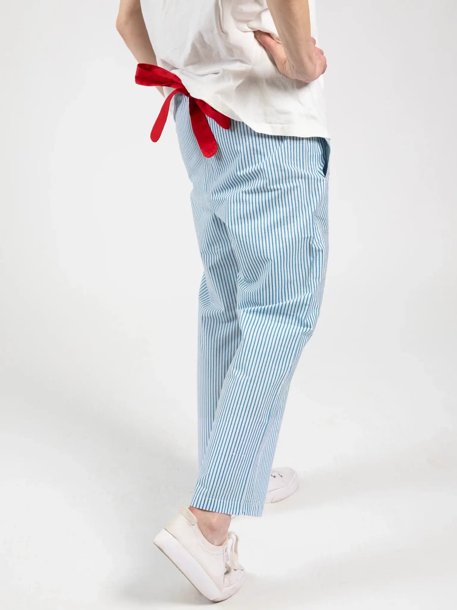 A woman wears Blue Stripe Women's Everyday Pants by Blue Stripe, featuring blue and white stripes and an elastic waistband, paired with white shoes and a white shirt with a red ribbon, standing against a plain white background.