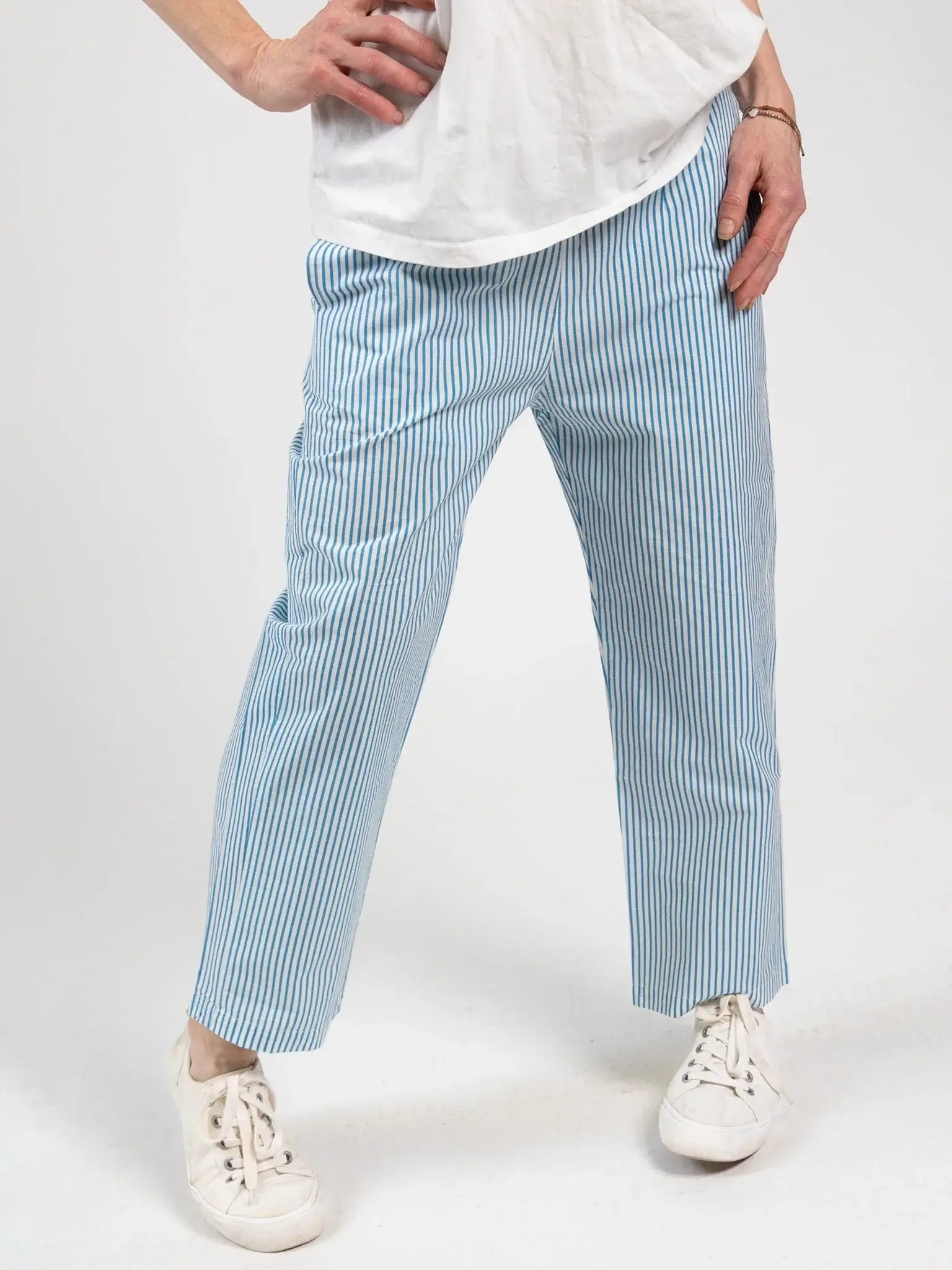 A person stands with one hand on hip, wearing a white T-shirt, Blue Stripe Women's Everyday Pants by Blue Stripe—ethically made blue and white striped cotton trousers with an elastic waistband—and white sneakers against a plain background.