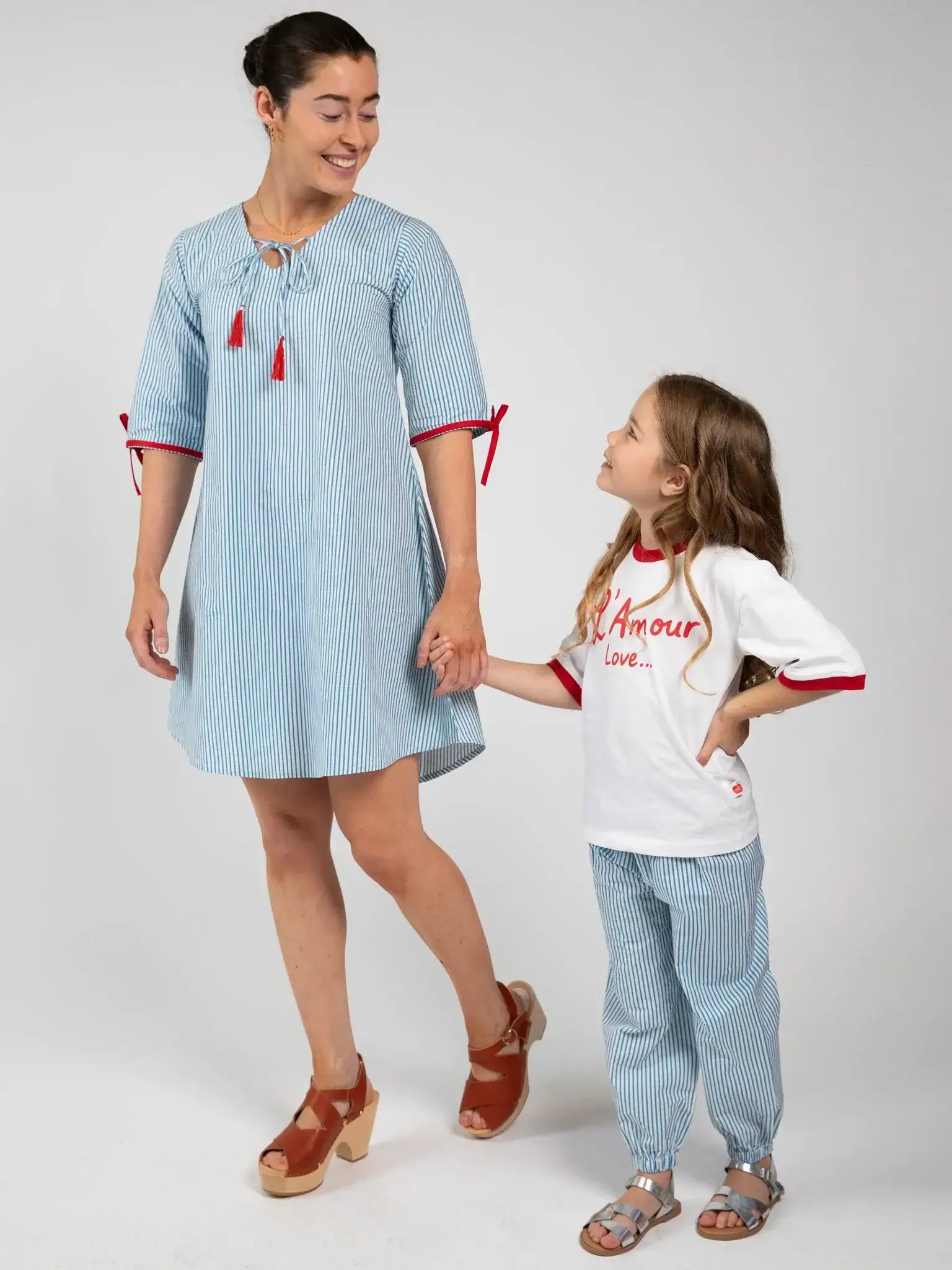 An adult and child hold hands, looking at each other. The adult wears the Blue Stripe Women's Harlow Kaftan Dress with brown sandals; the child wears a t-shirt, striped pants, and sandals.