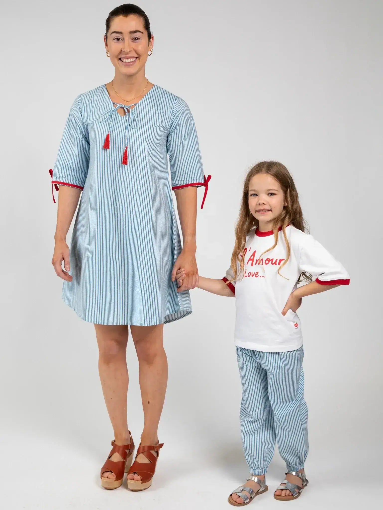 An adult in a Blue Stripe Women's Harlow Kaftan Dress and sandals holds hands with a child wearing a white shirt with red text, 100% cotton blue-striped pants, and sandals.