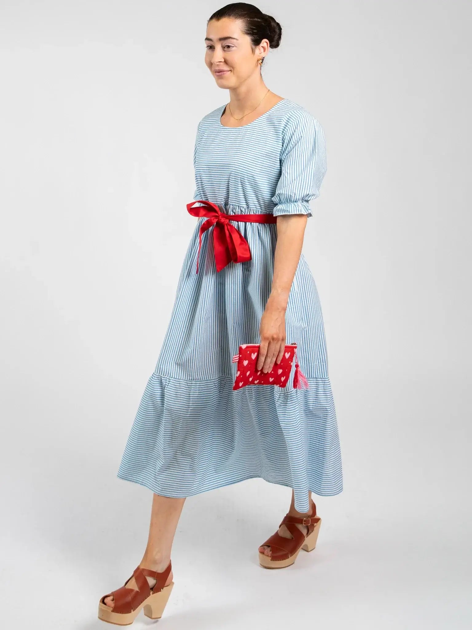 A woman in the Blue Stripe Women’s Blue Stripe Maxi Dress, featuring ethical design, pairs it with a red ribbon belt, red polka dot clutch, and brown platform sandals against a plain background.