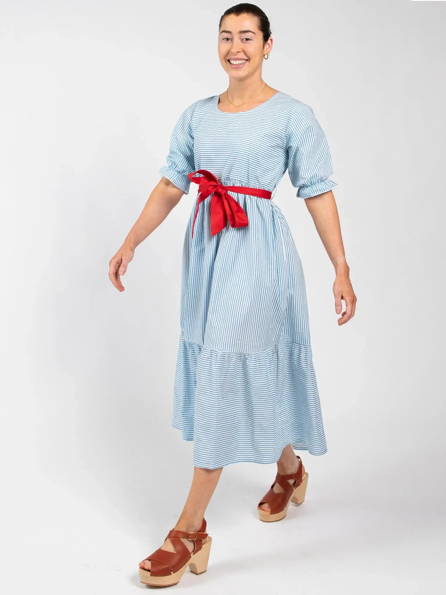 A woman in the Blue Stripe Women's Blue Stripe Maxi Dress with puff sleeves and a red waist ribbon, paired with tan platform sandals, walks against a white background.