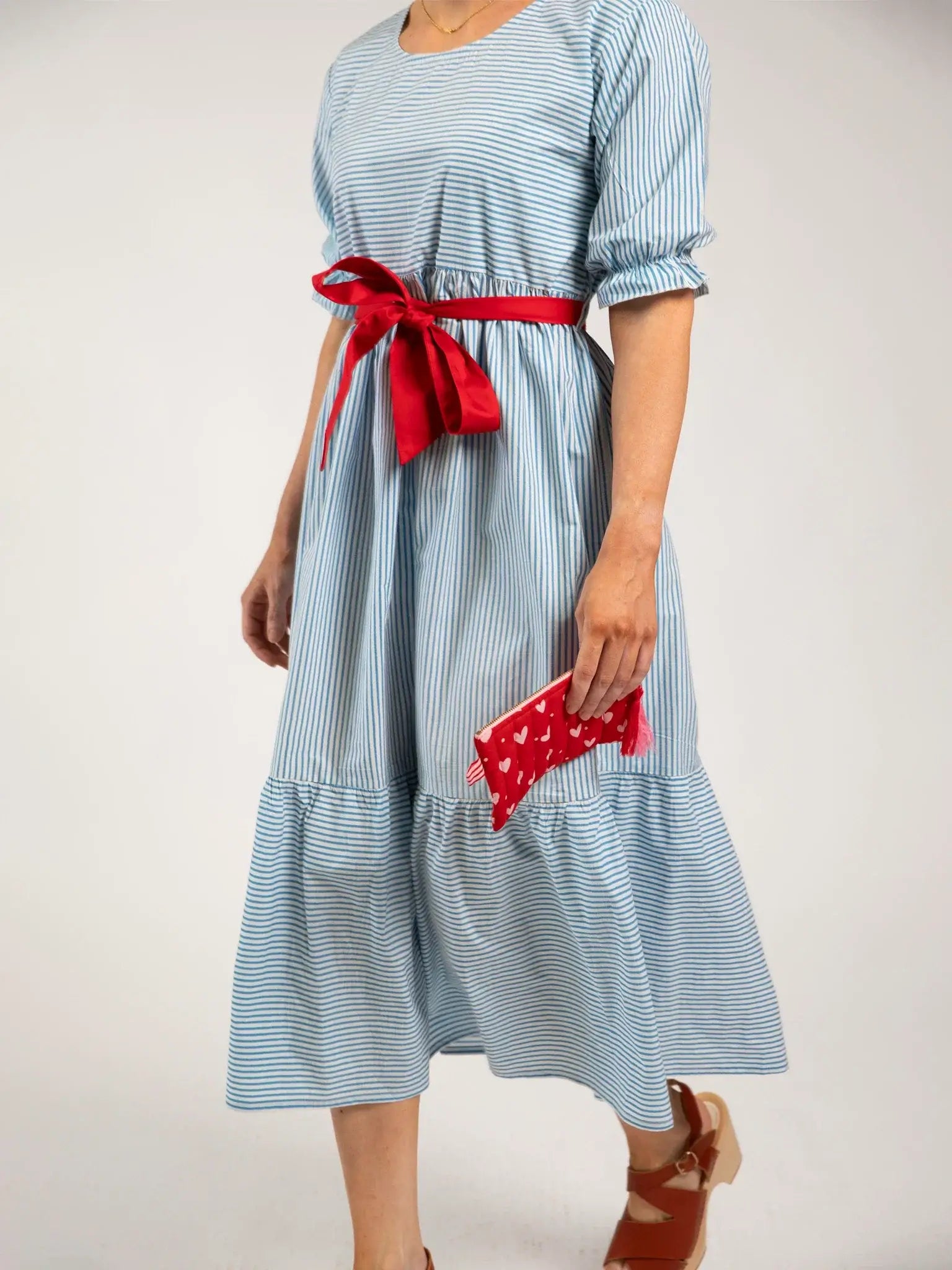 A woman in a Blue Stripe brand Women's Blue Stripe Maxi Dress with a red ribbon belt, holding a red clutch and wearing brown sandals.