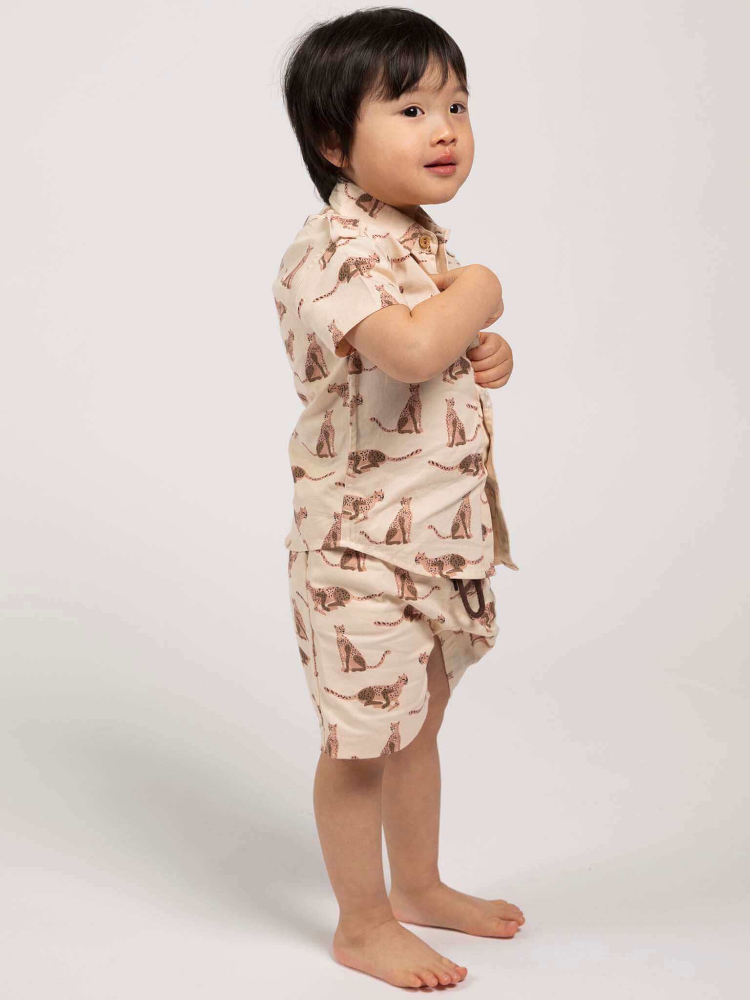 Child wearing a beige, leopard animal-patterned collared shirt and shorts with plain background
