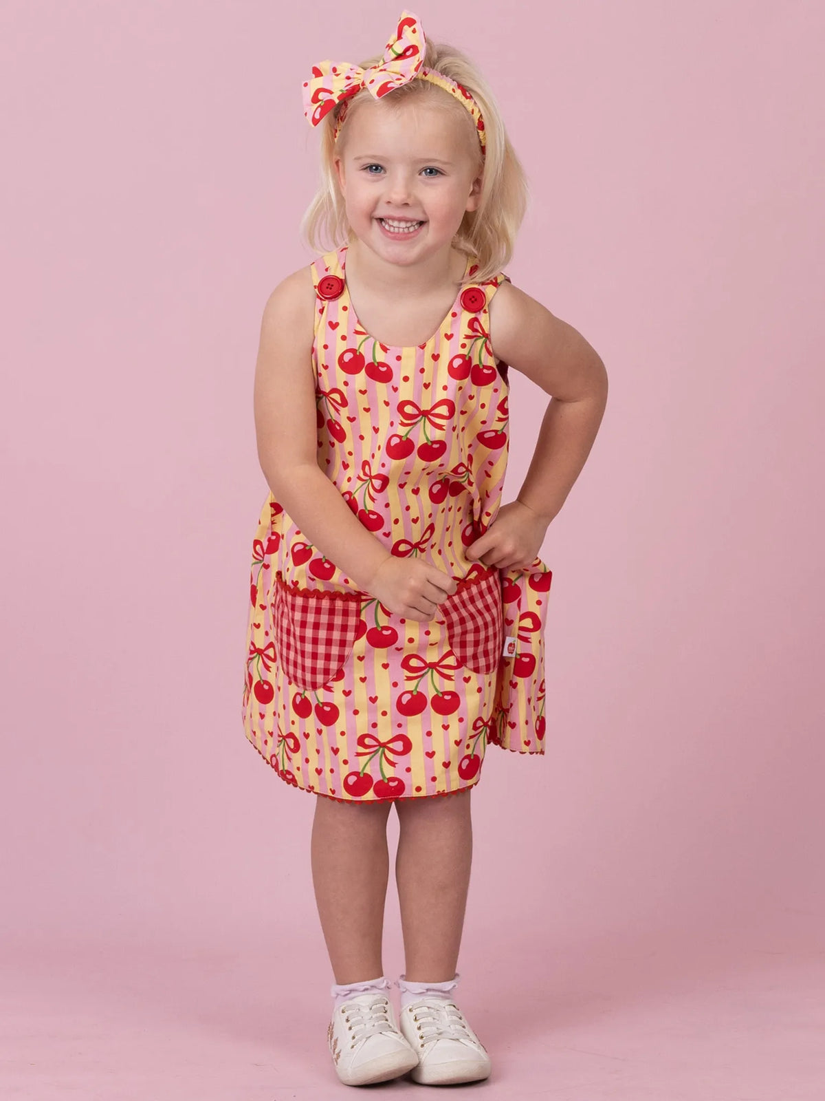 A young girl with blonde hair wearing a red and yellow cherry print sleeveless dress with a matching headband, standing against a solid pink background.