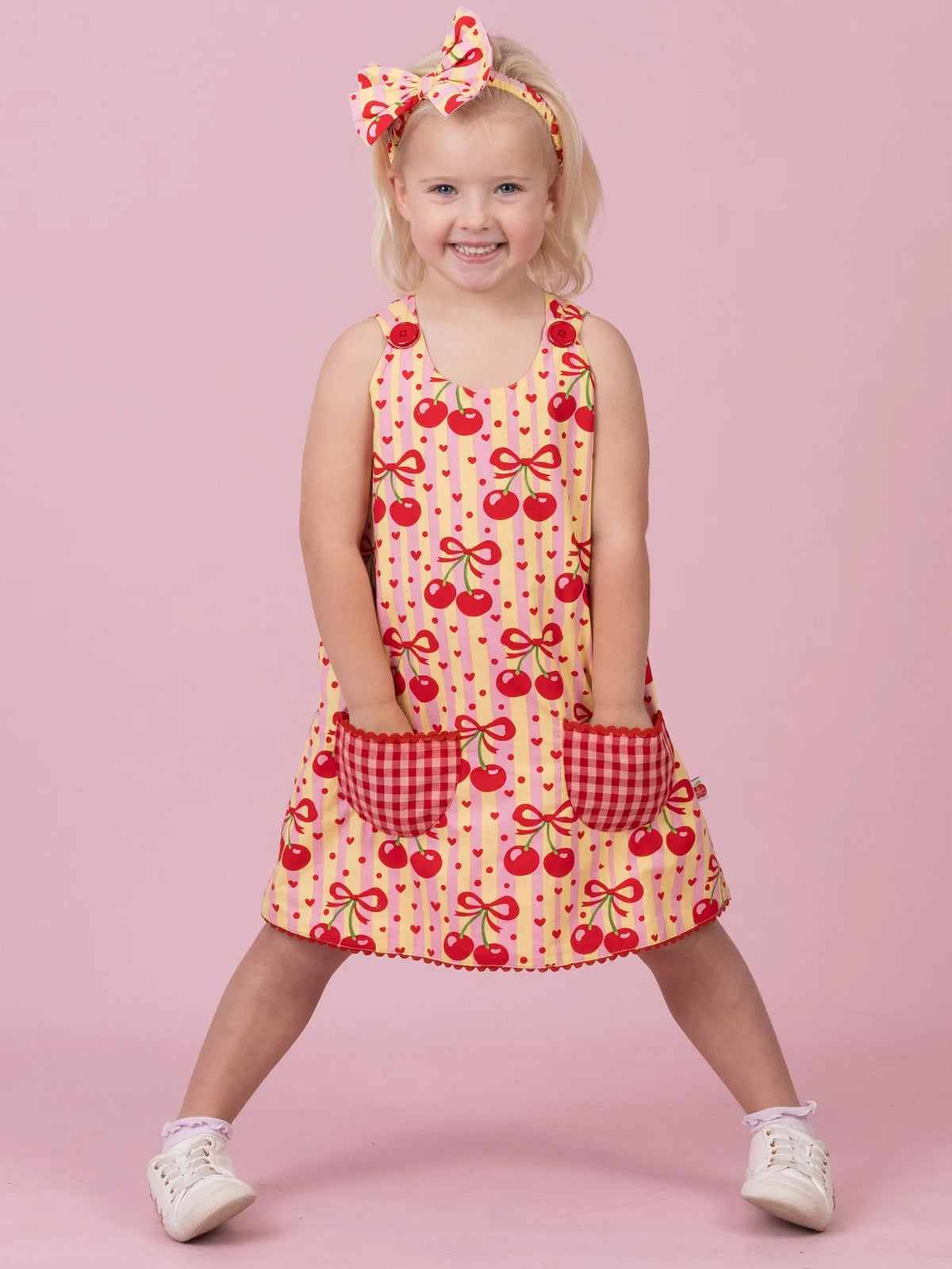 A young girl stands against a solid pink studio background wearing a sleeveless yellow dress with a red cherry print, red gingham pockets, and a matching red-and-yellow bow headband.