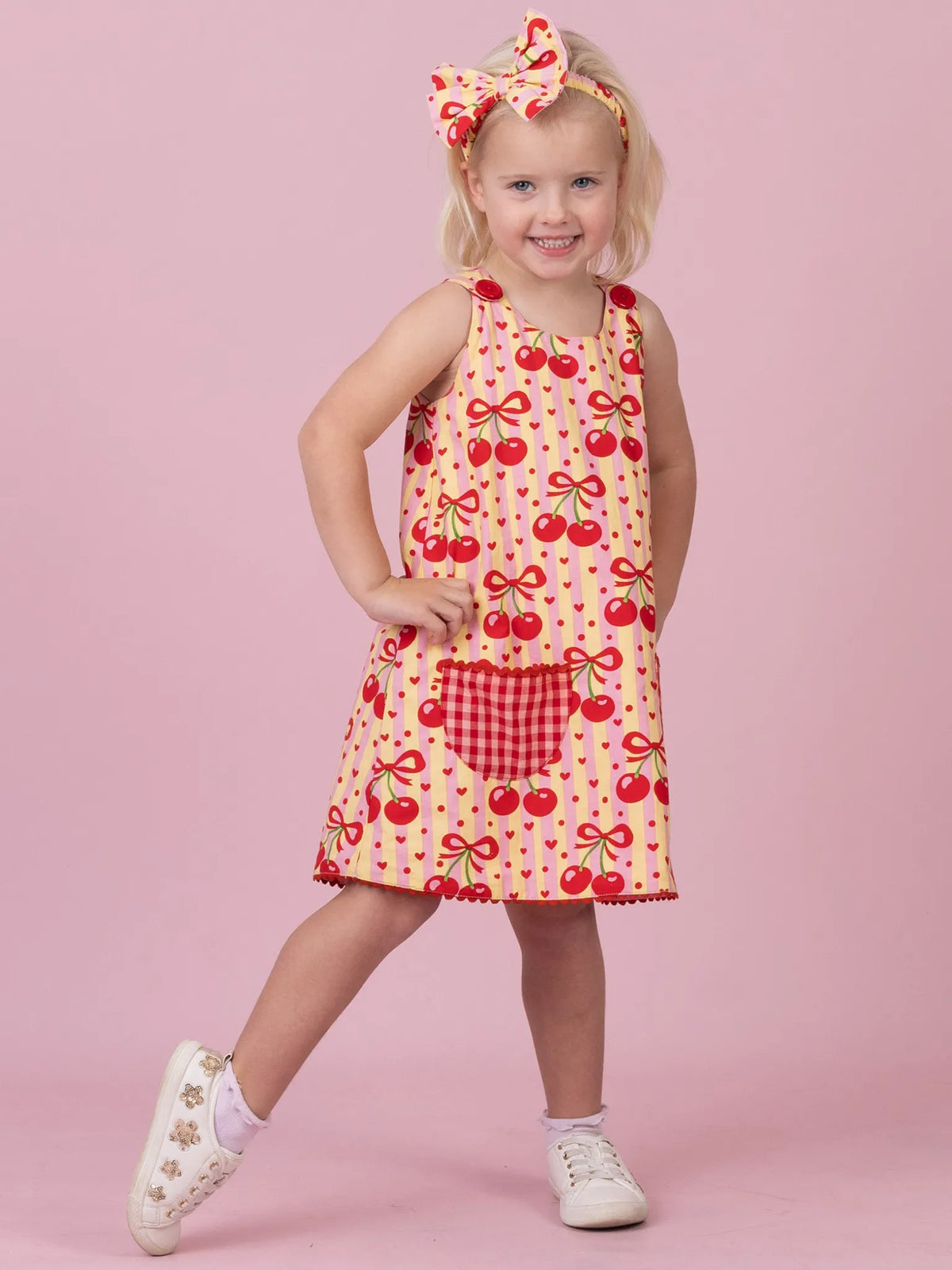 A young girl modeling a sleeveless red and yellow cherry print dress with a gingham pocket, matching headband, and white sneakers, set against a solid pink studio background.