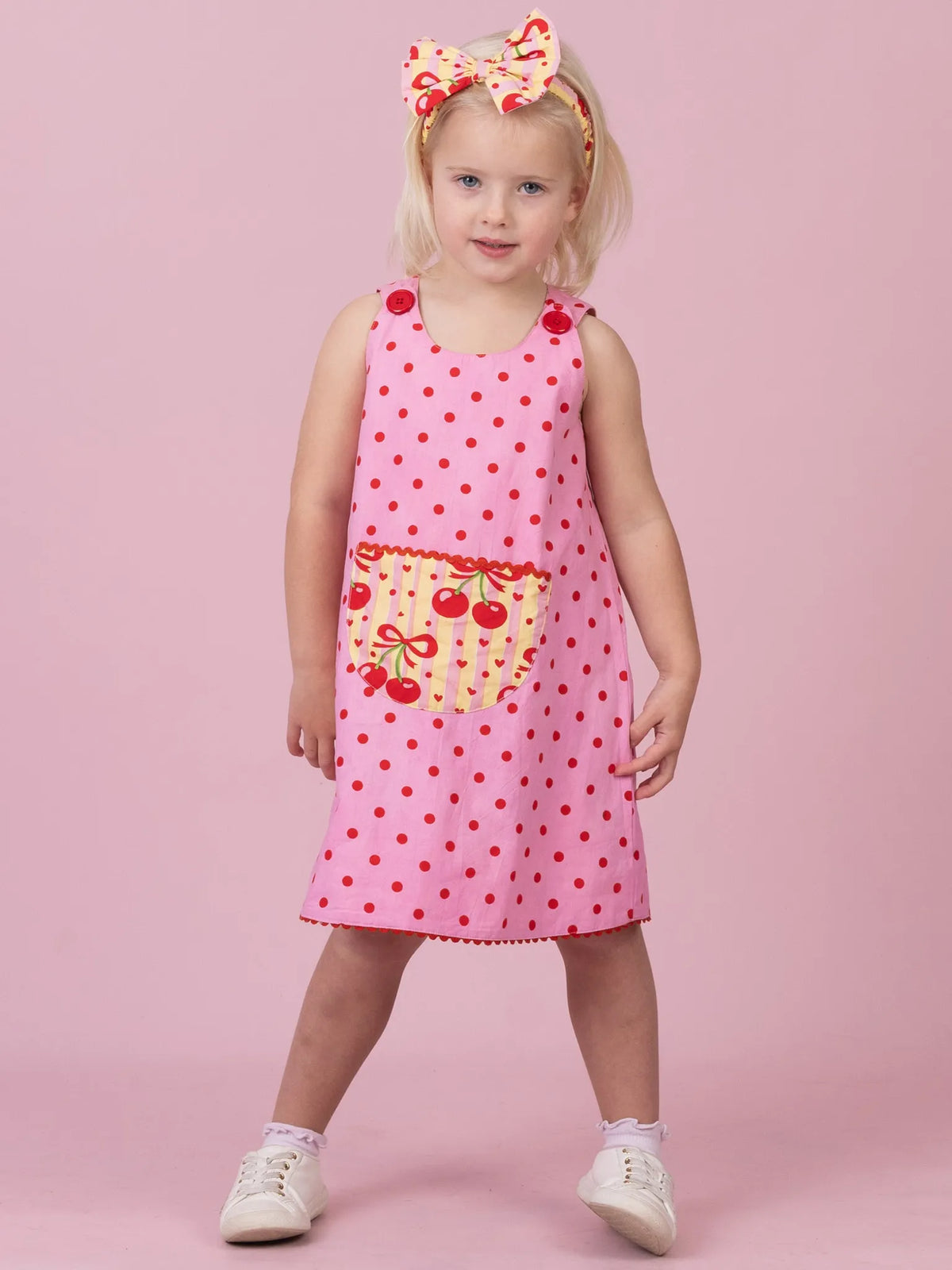 A young girl stands against a solid pink studio background wearing a pink sleeveless dress with red polka dots and a cherry-themed front pocket, along with a matching headband and white sneakers.