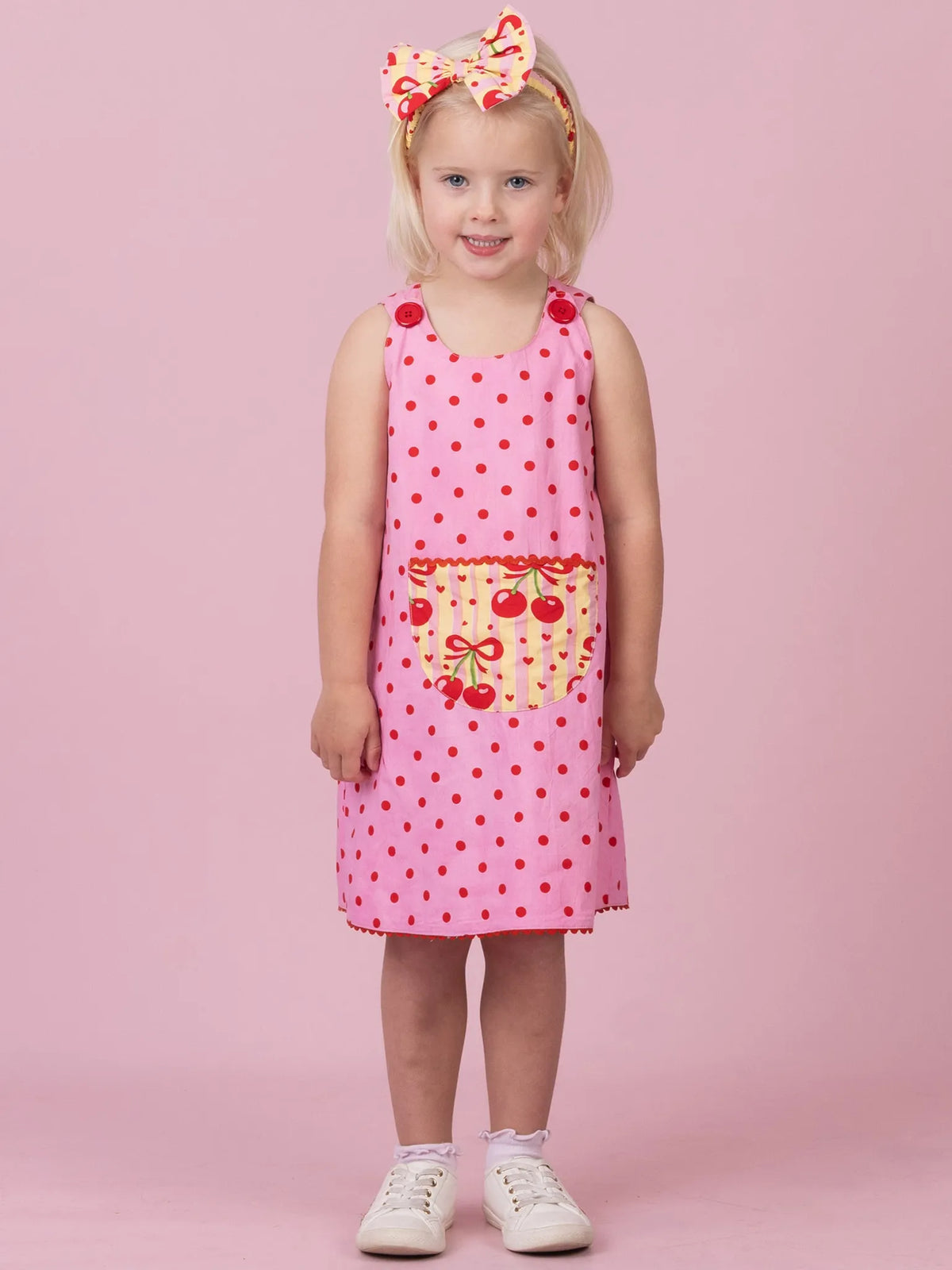 A young girl stands against a solid pink studio background wearing a pink sleeveless dress with red polka dots, a cherry-patterned front pocket, a matching pink bow headband, and white sneakers.