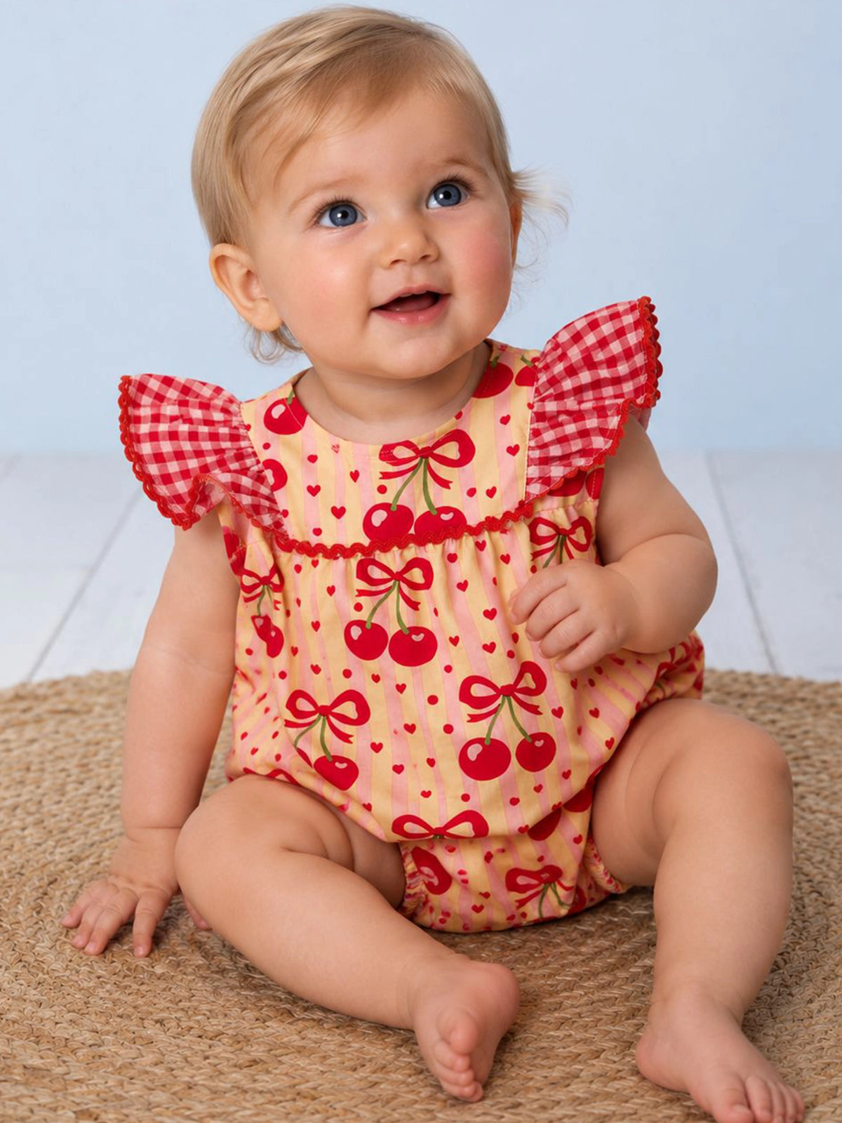 Baby wearing a red and yellow romper with cherry pattern on a light blue background