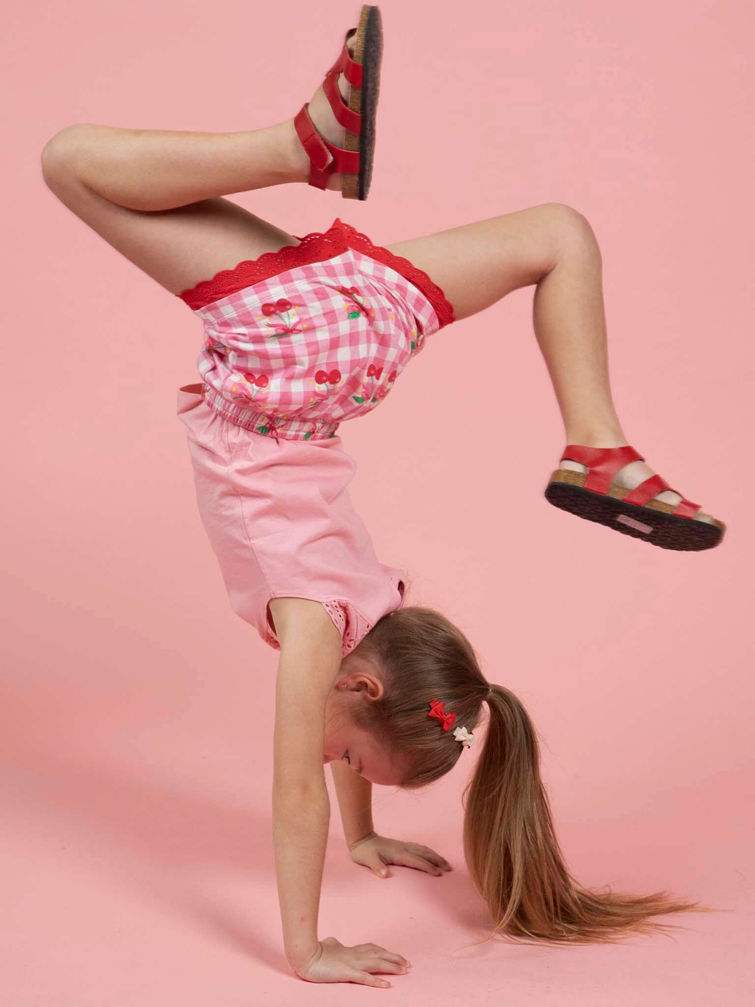 A young girl in Cherry Gingham Jersey Shorts and red sandals does a handstand against a pink background.