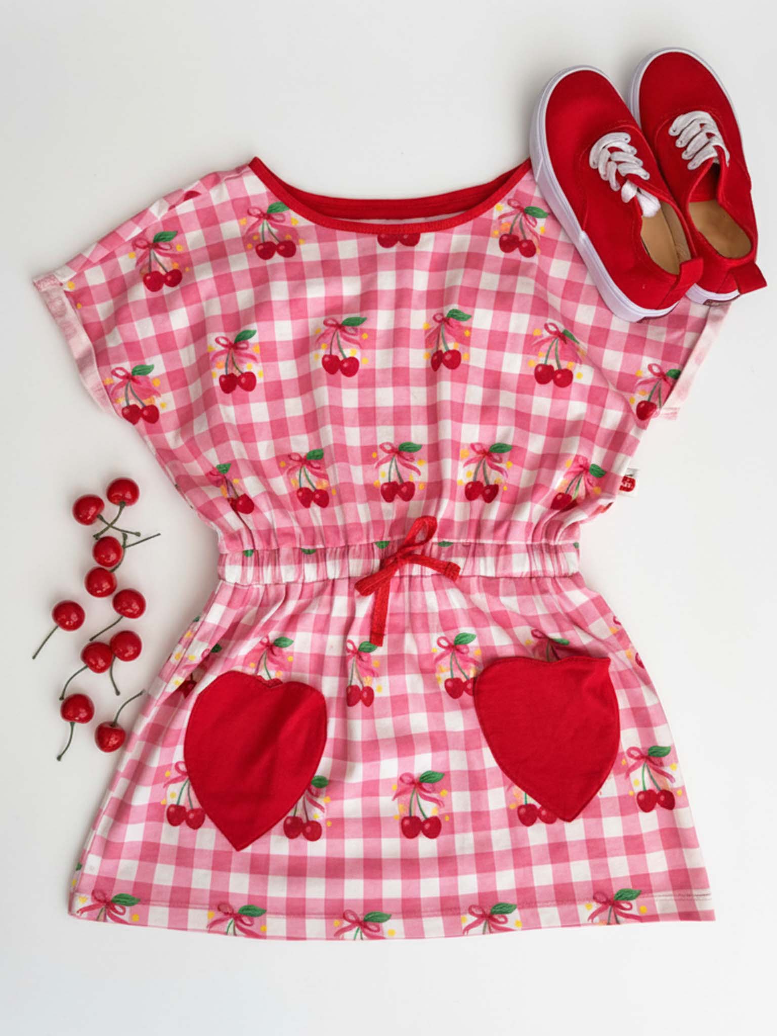 Flat lay of the New Love Cherry Gingham Jersey Dress by Cherry Gingham, featuring a pink and white gingham cherry print, red heart-shaped pockets, an elastic waist, styled with red shoes and cherries beside this girls' cotton dress.