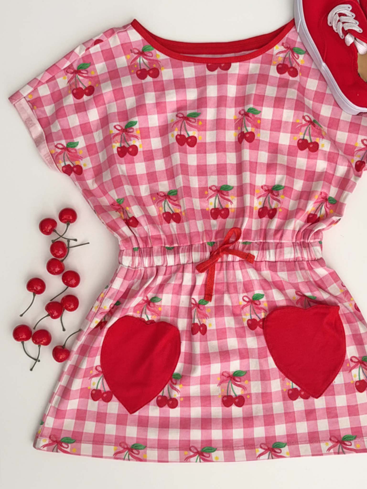 The Cherry Gingham New Love Cherry Gingham Jersey Dress features pink and white gingham, cherry print, red heart pockets, a red waistband, artificial cherries, and is perfect for playful days.