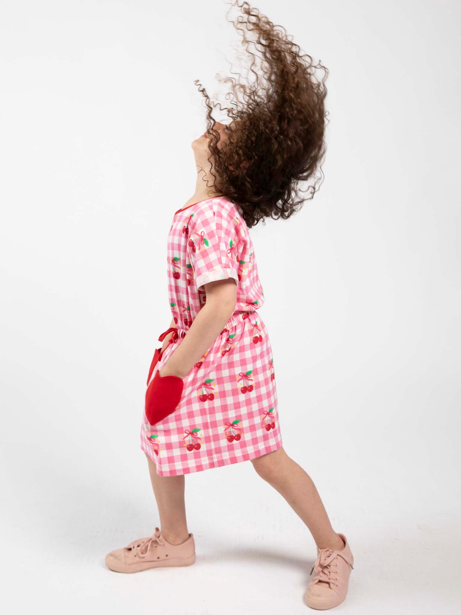 A young girl wears the Cherry Gingham New Love Cherry Gingham Jersey Dress with cherry patterns, an elastic waist, and pairs it with pink sneakers as she tilts her head back and flips her hair.