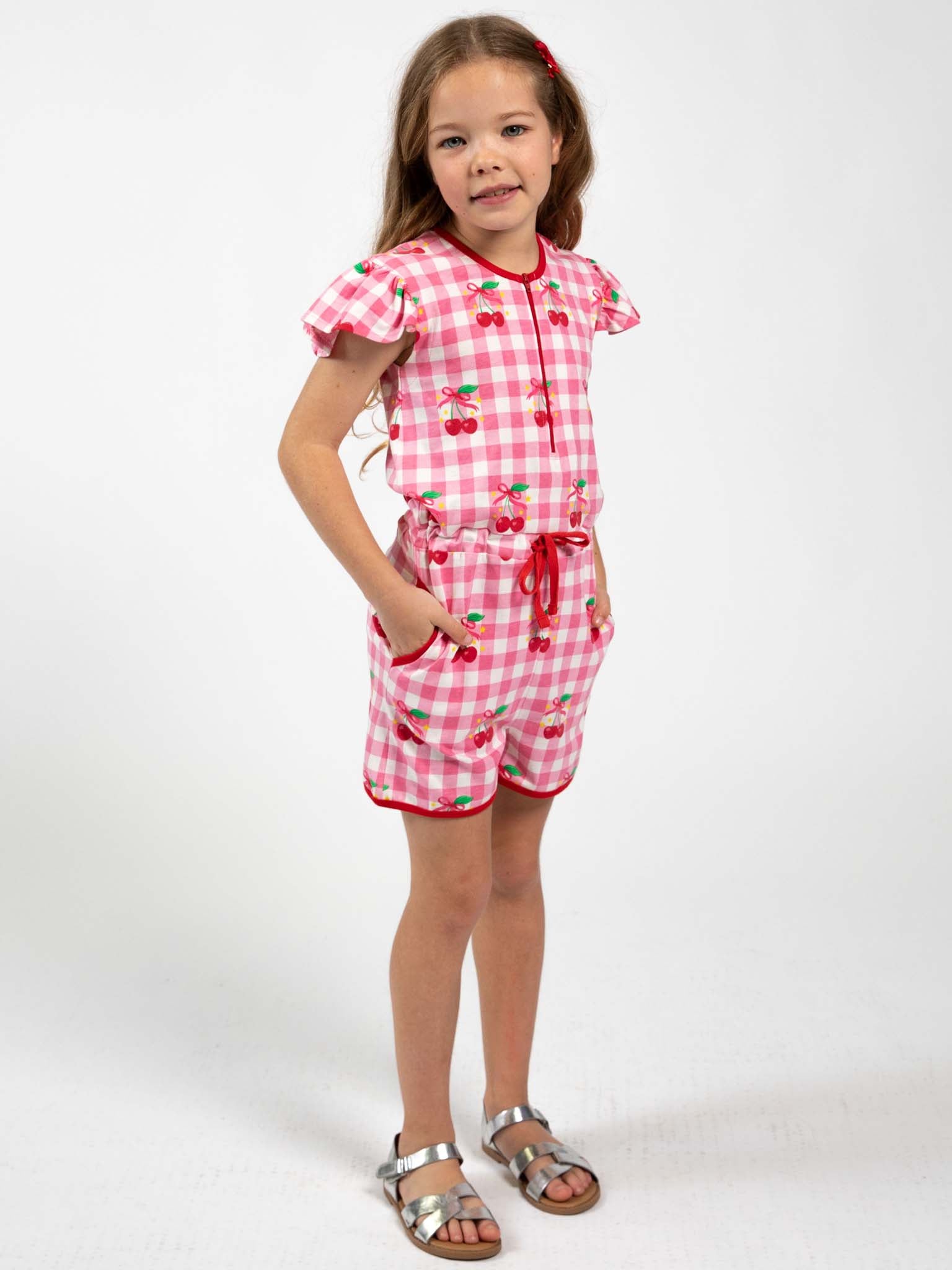 A young girl wears the Cherry Gingham Jersey Playsuit by Cherry Gingham, featuring cherry prints and gingham pattern, paired with silver sandals and a red hair clip against a plain white background.