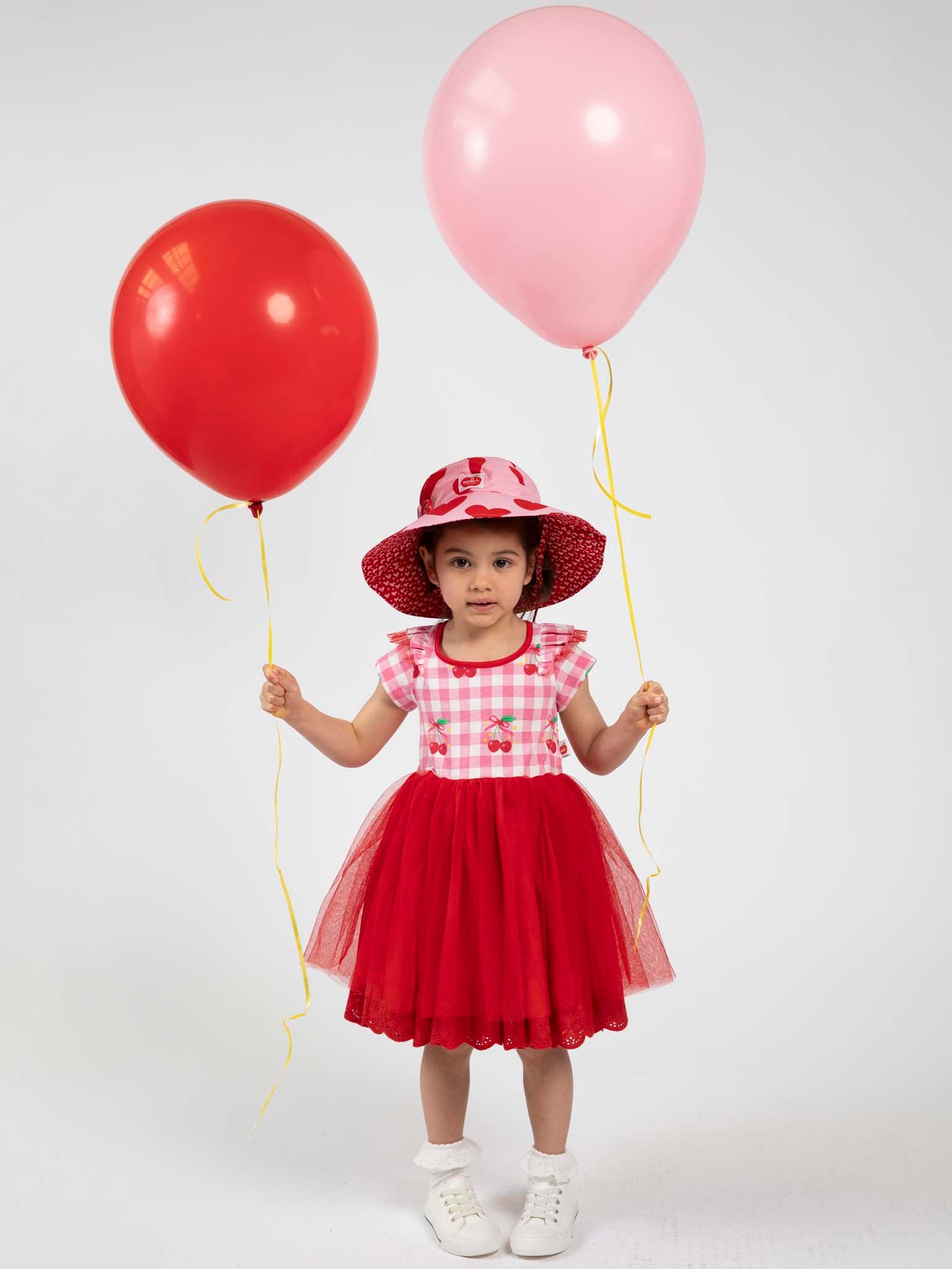 A young girl wears the Cherry Gingham Tutu Dress by Cherry Gingham, along with a matching hat, holding a red balloon in her right hand and a pink balloon in her left, standing against a plain white background.
