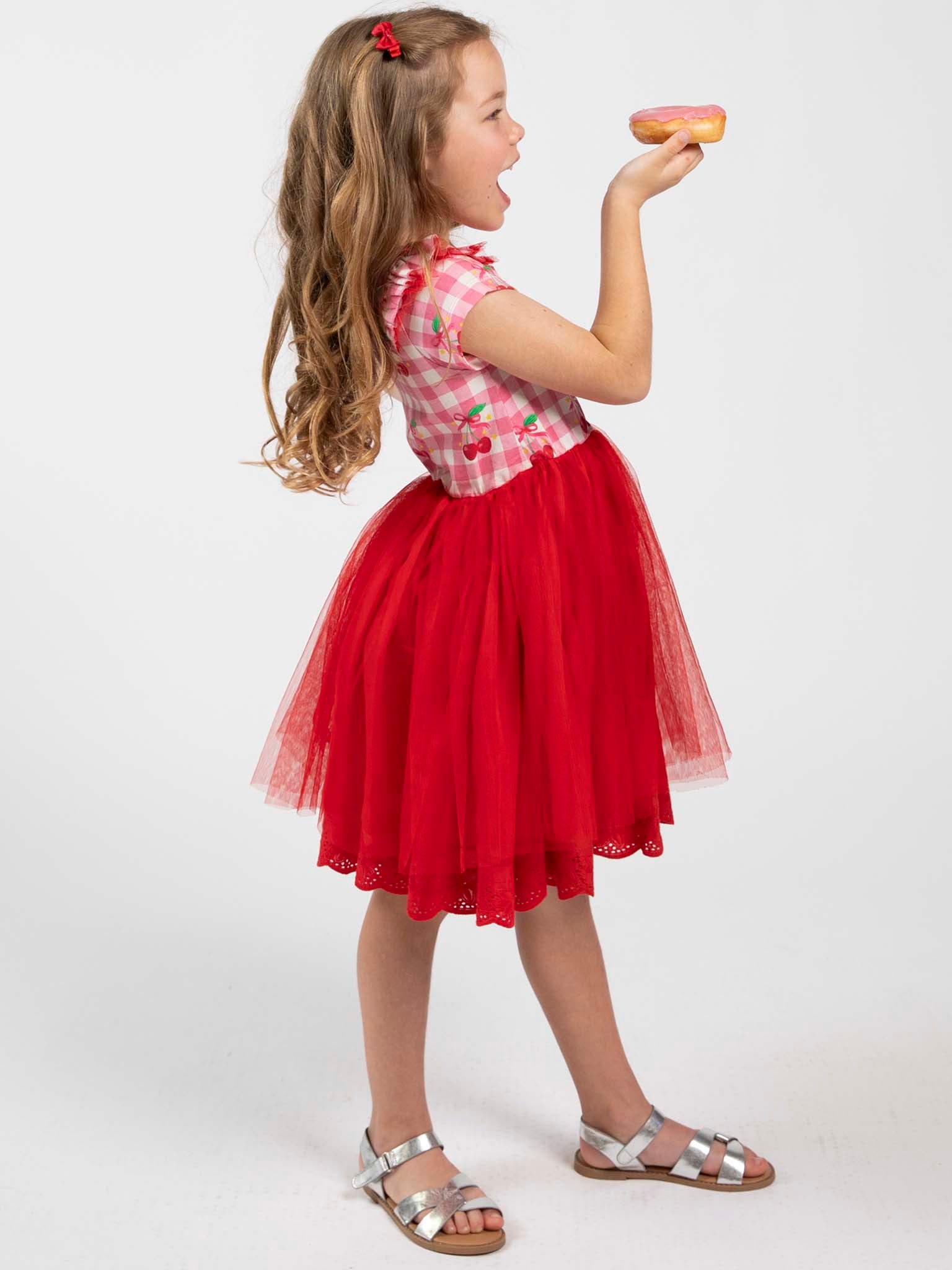 A young girl in the Cherry Gingham Tutu Dress by Cherry Gingham stands sideways, holding a doughnut near her mouth and gazing at it.