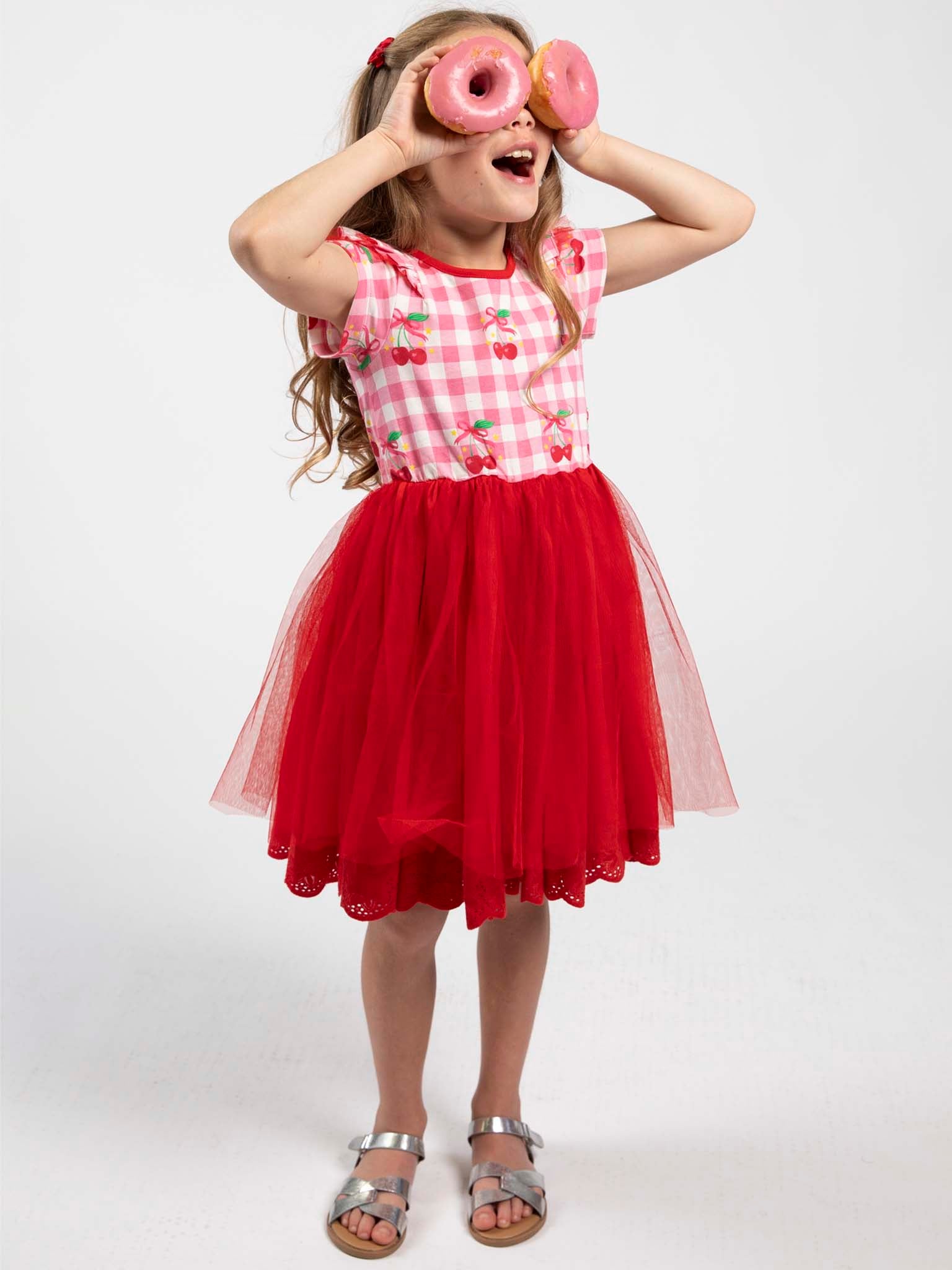 A young girl wears the Cherry Gingham Tutu Dress by Cherry Gingham, playfully holding two pink donuts over her eyes against a white background for a charming vintage vibe.