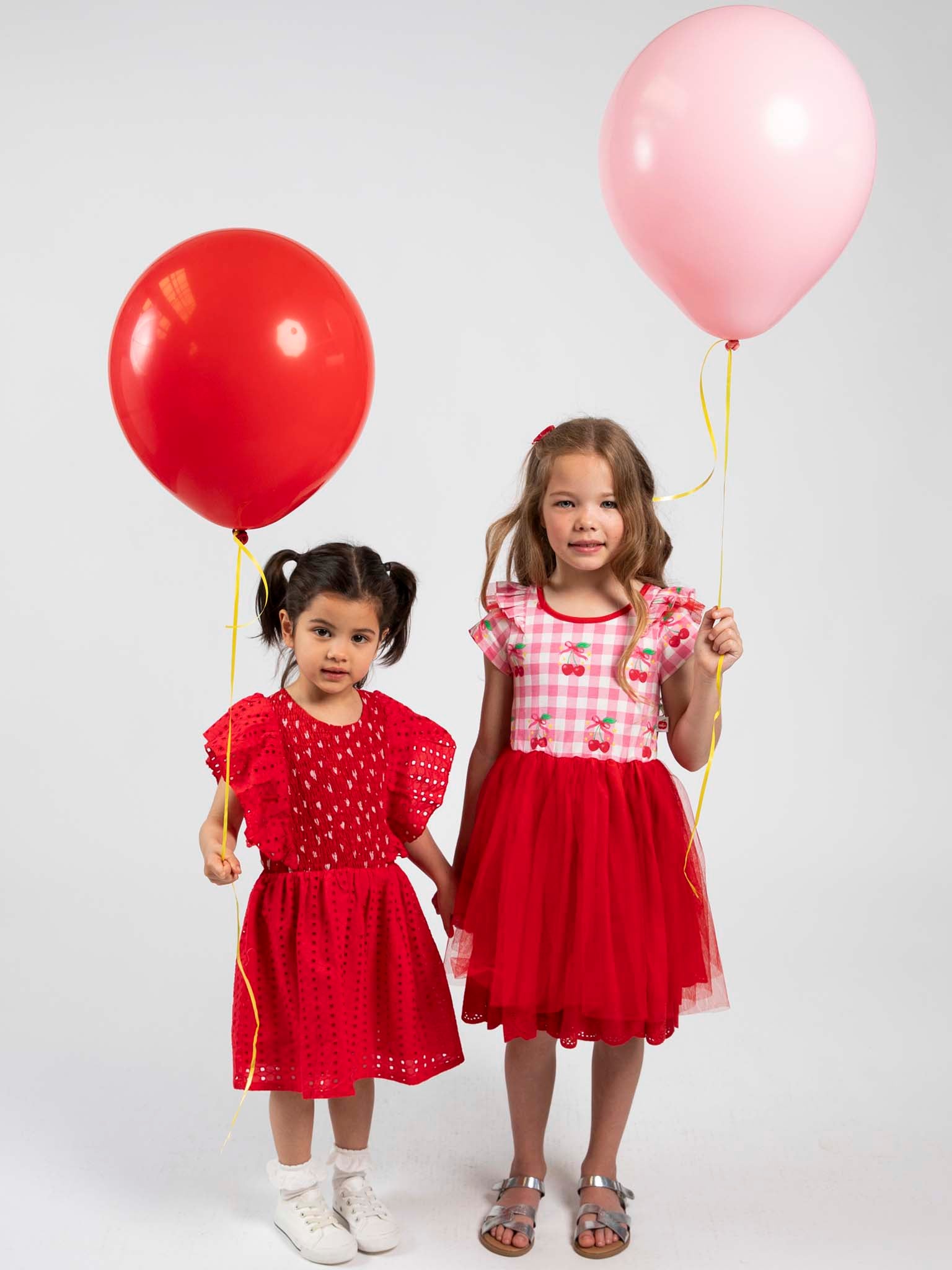 Two young girls in red dresses holding red and pink balloons against a white background