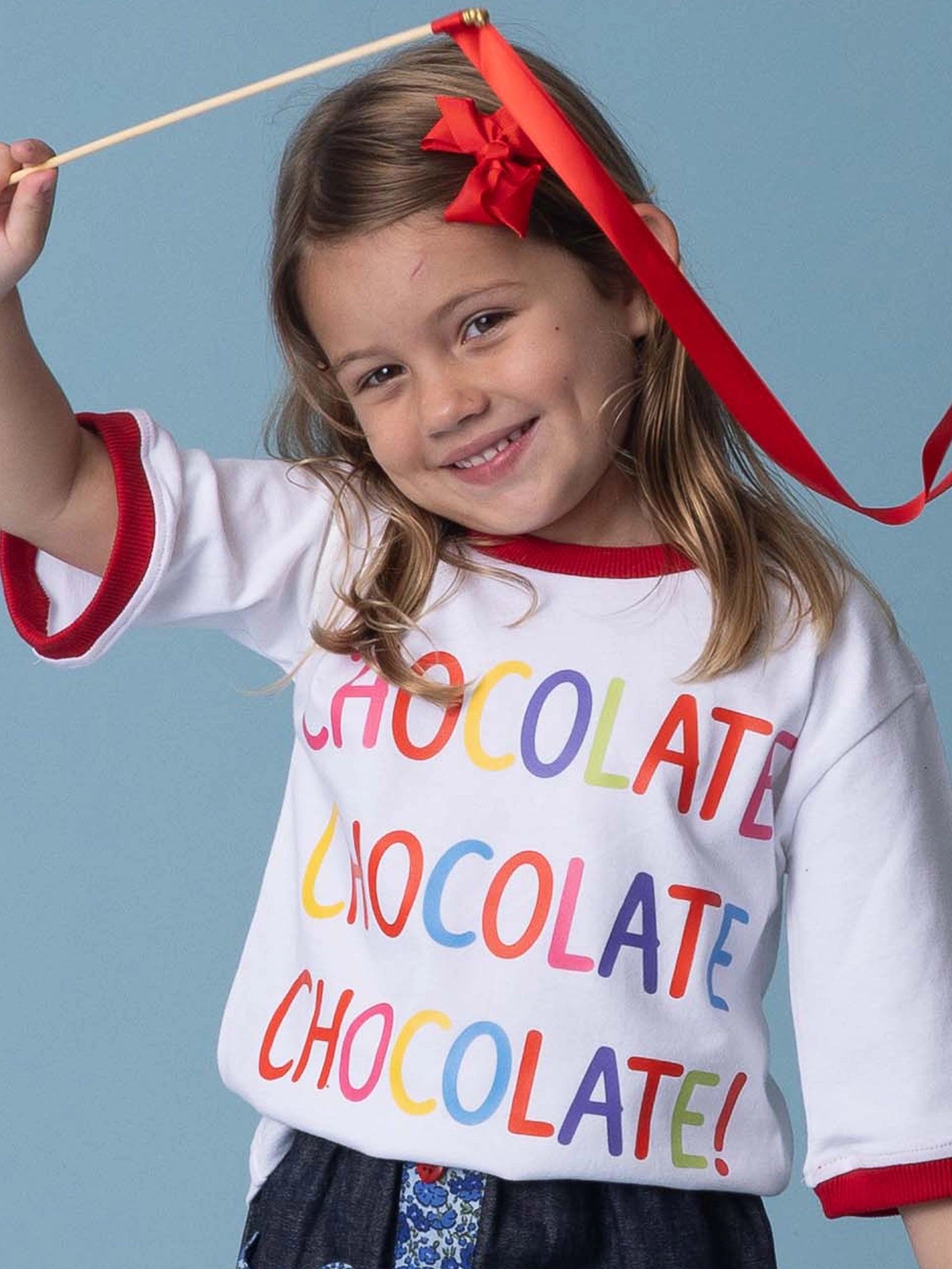 A smiling young girl holds a red ribbon and wears the Essentials Chocolate Tee with Red Binding—personalise it!.