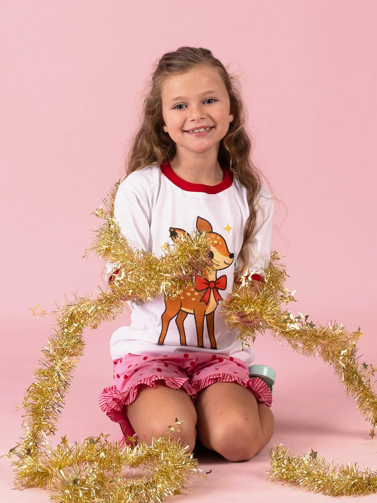 Girl kneeling in gold tinsel in the white Christmas Deer tee and Pink & Red Dots shorts
