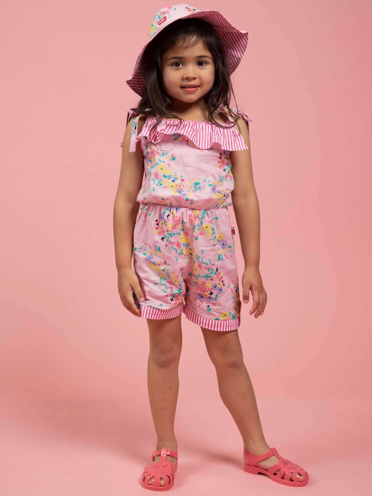 A young girl wears the Confetti Floral Josie Playsuit by Confetti Floral with a matching summer hat and pink sandals, standing against a solid pink background.
