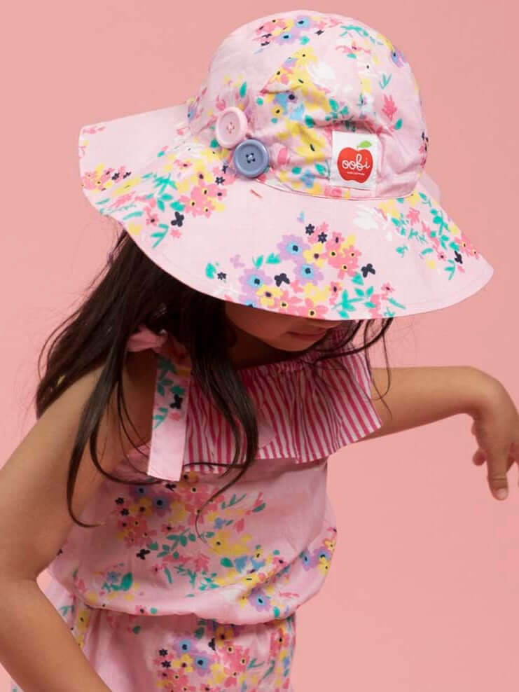A young girl wearing a Confetti Floral Ponytail Hat and matching floral dress stands against a pink background.