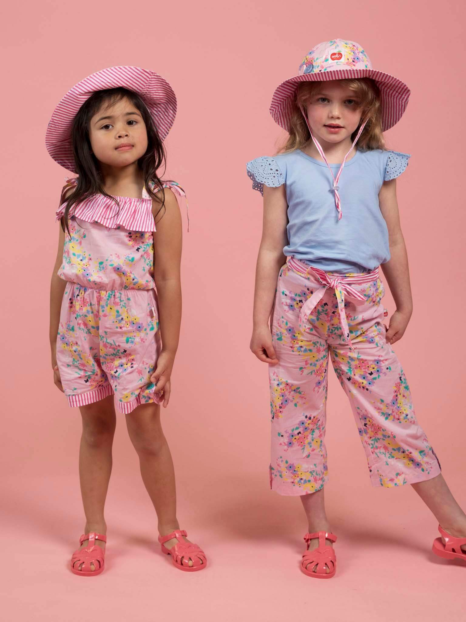 Two young girls stand against a pink background in colorful summer outfits and sandals, each wearing a Confetti Floral Ponytail Hat by Confetti Floral for the perfect adjustable fit.