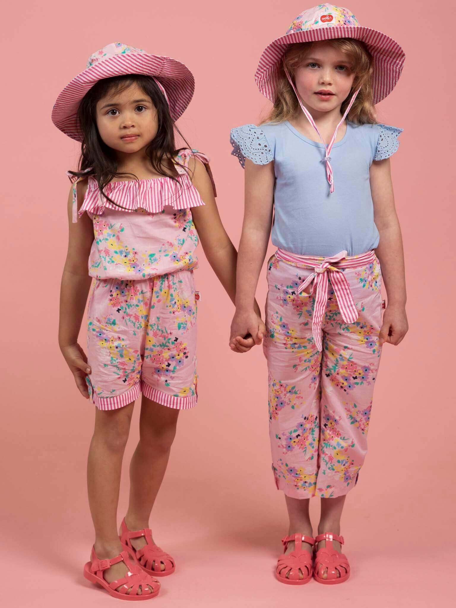 Two young girls stand hand-in-hand before a pink backdrop, dressed in floral outfits and pink sandals. Their Confetti Floral Ponytail Hats offer both style and sun protection—ideal for playful days.