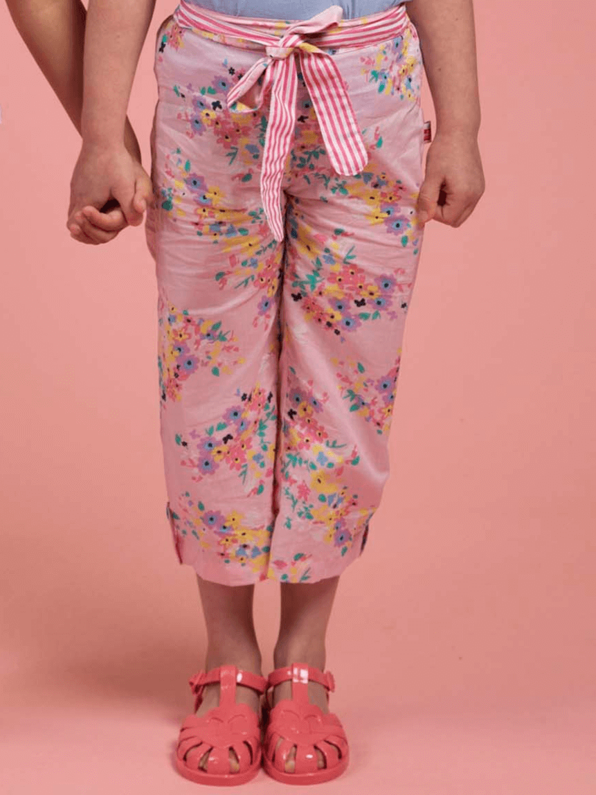 Two children hold hands; one wears Confetti Floral Wide-Leg Pants by Confetti Floral, paired with a striped belt and pink sandals, set against a pink background.
