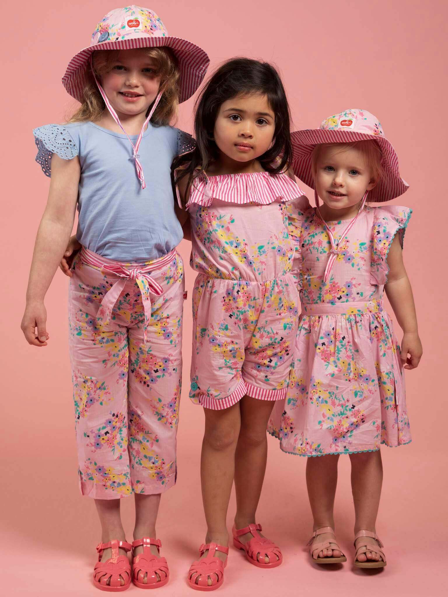 Three young girls pose before a pink backdrop in coordinated Confetti Floral wide-leg pants and hand-printed pastel outfits, paired with pink sandals; two of them also wear matching girls’ hats.