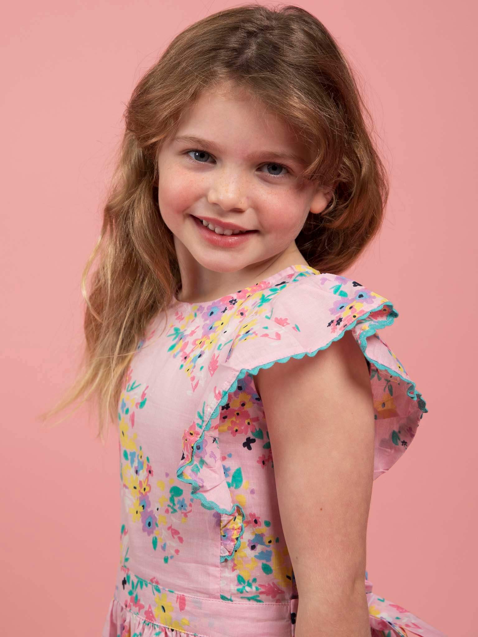 A young girl with long, light brown hair smiles at the camera against a pink background, wearing the Confetti Floral Lacy Dress by Confetti Floral.