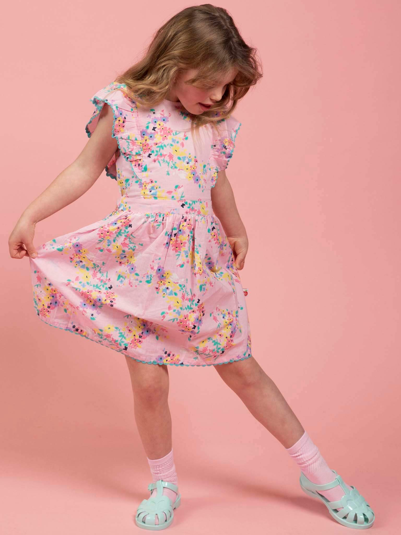 A young girl in the Confetti Floral Lacy Dress by Confetti Floral stands against a pink background, looking down and holding out the side of her dress.