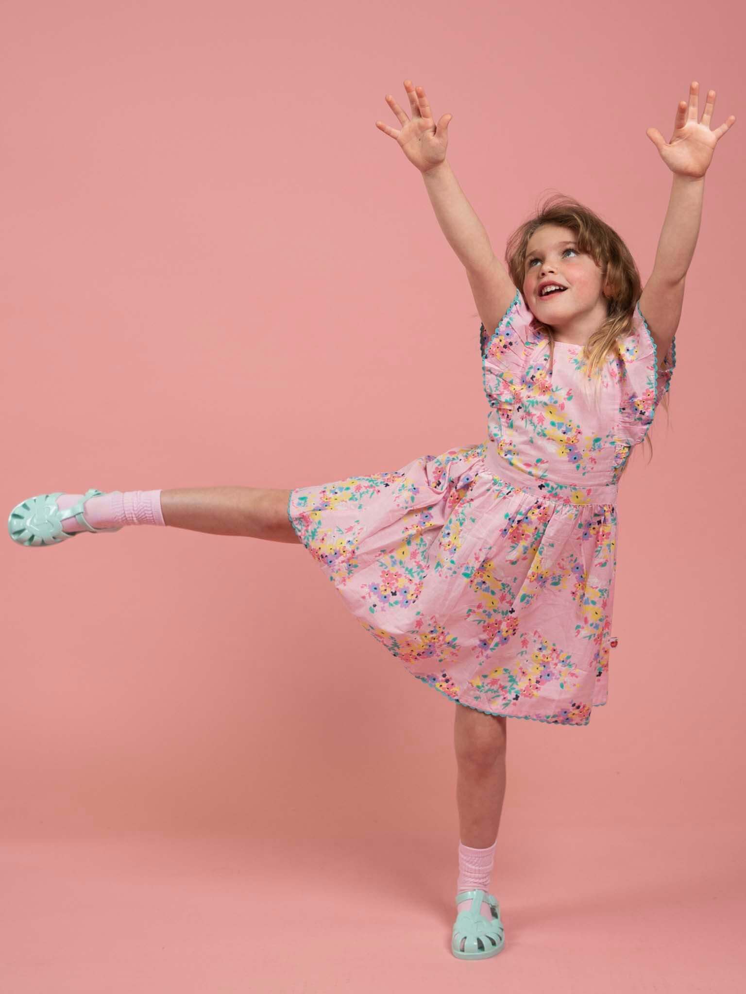 A young girl poses on one leg with arms raised in the Confetti Floral Lacy Dress by Confetti Floral, styled with teal shoes against a plain pink background.