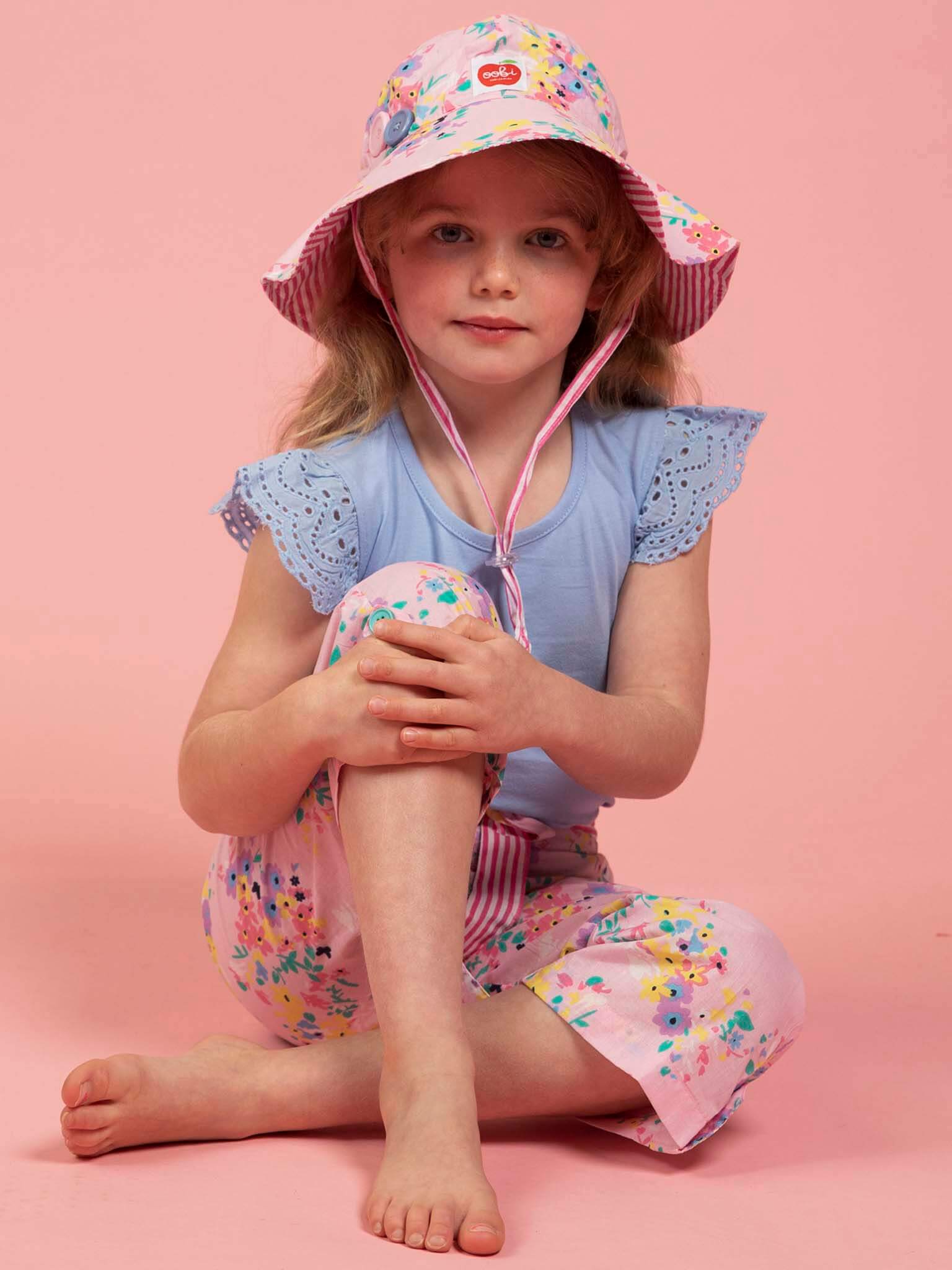 A young girl sits cross-legged on a pink background, wearing Confetti Floral Wide-Leg Pants by Confetti Floral, a floral sun hat from our girls hats collection, and a blue top with eyelet sleeves.