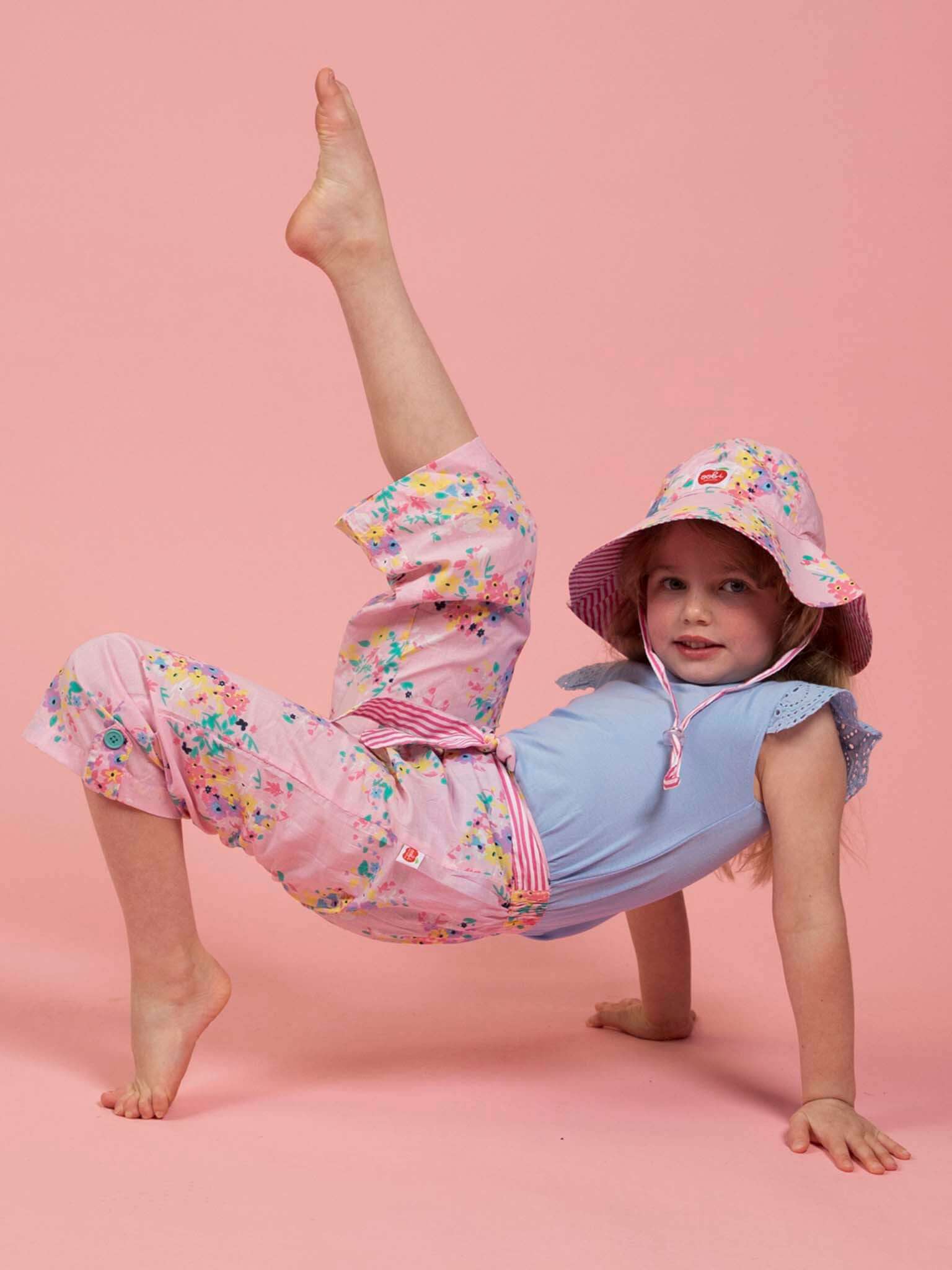 A young girl poses on the floor with one leg raised, wearing Confetti Floral Wide-Leg Pants by Confetti Floral, a floral hat, and a blue top against a pink background.