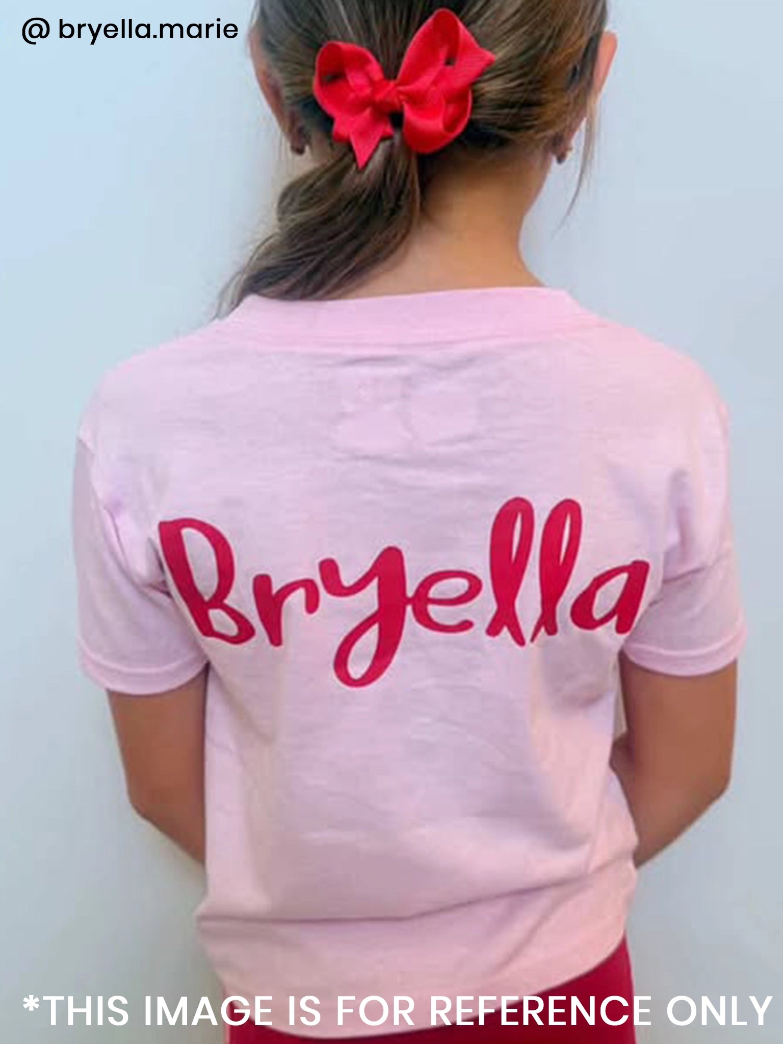 A girl with a red bow wears the Essentials Deer Printed Tee, available in sizes 2Y to adult, made from 100% cotton in an oversized light pink fit, personalised with "Bryella" in bold red letters on the back.