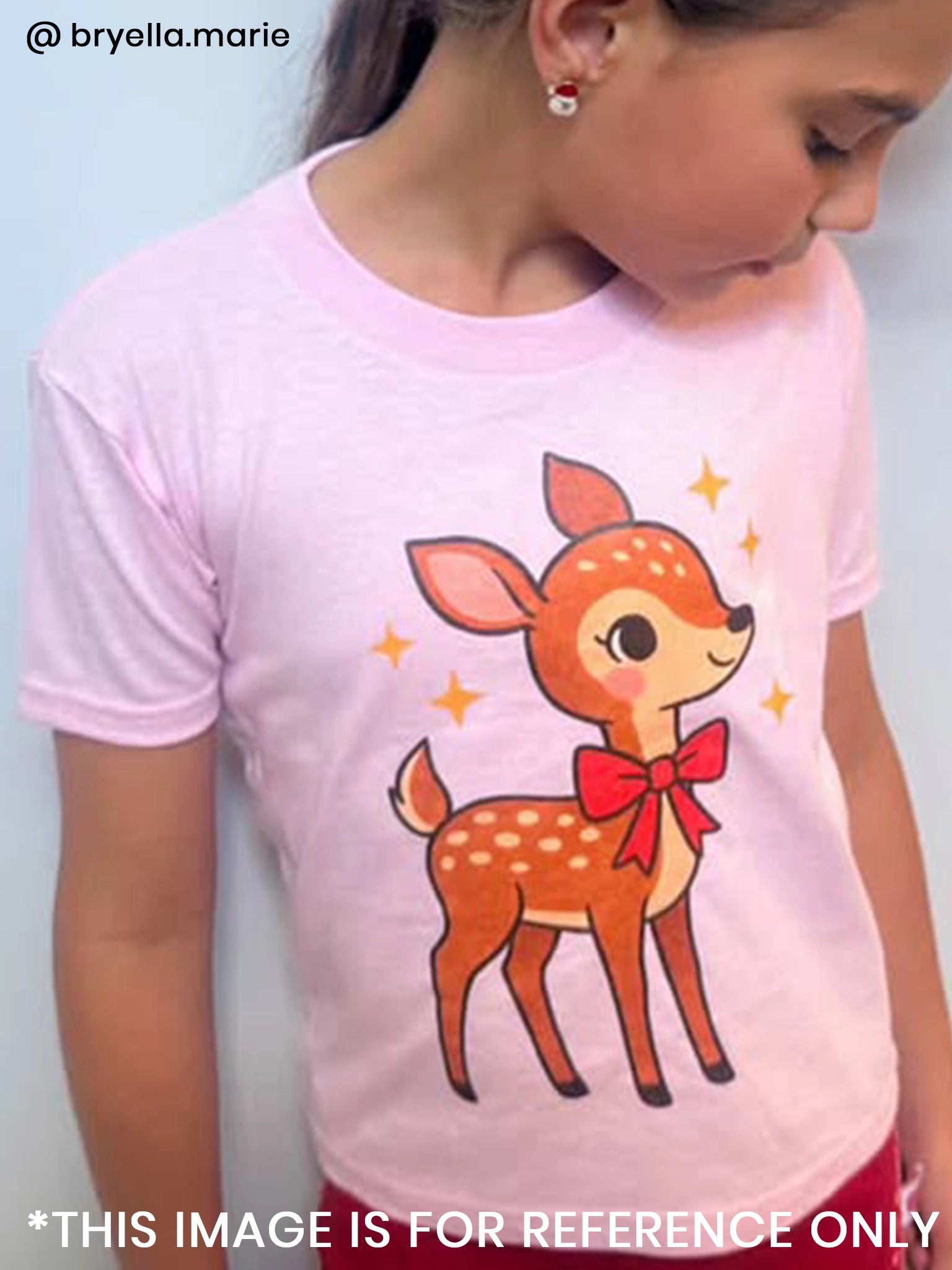 A girl wears the Essentials Deer Printed Tee—an oversized, light pink shirt with a cartoon fawn, red bow, and sparkles on the front. Personalisation available; sizes from 2Y to adult.
