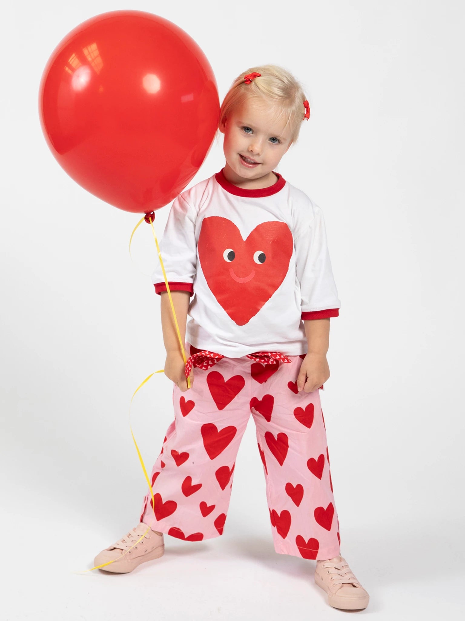 A young child stands against a plain white background, holding a red balloon and wearing Big Hearts' Big Heart Wide-Leg Pants with red heart prints, paired with a white shirt featuring a heart design.