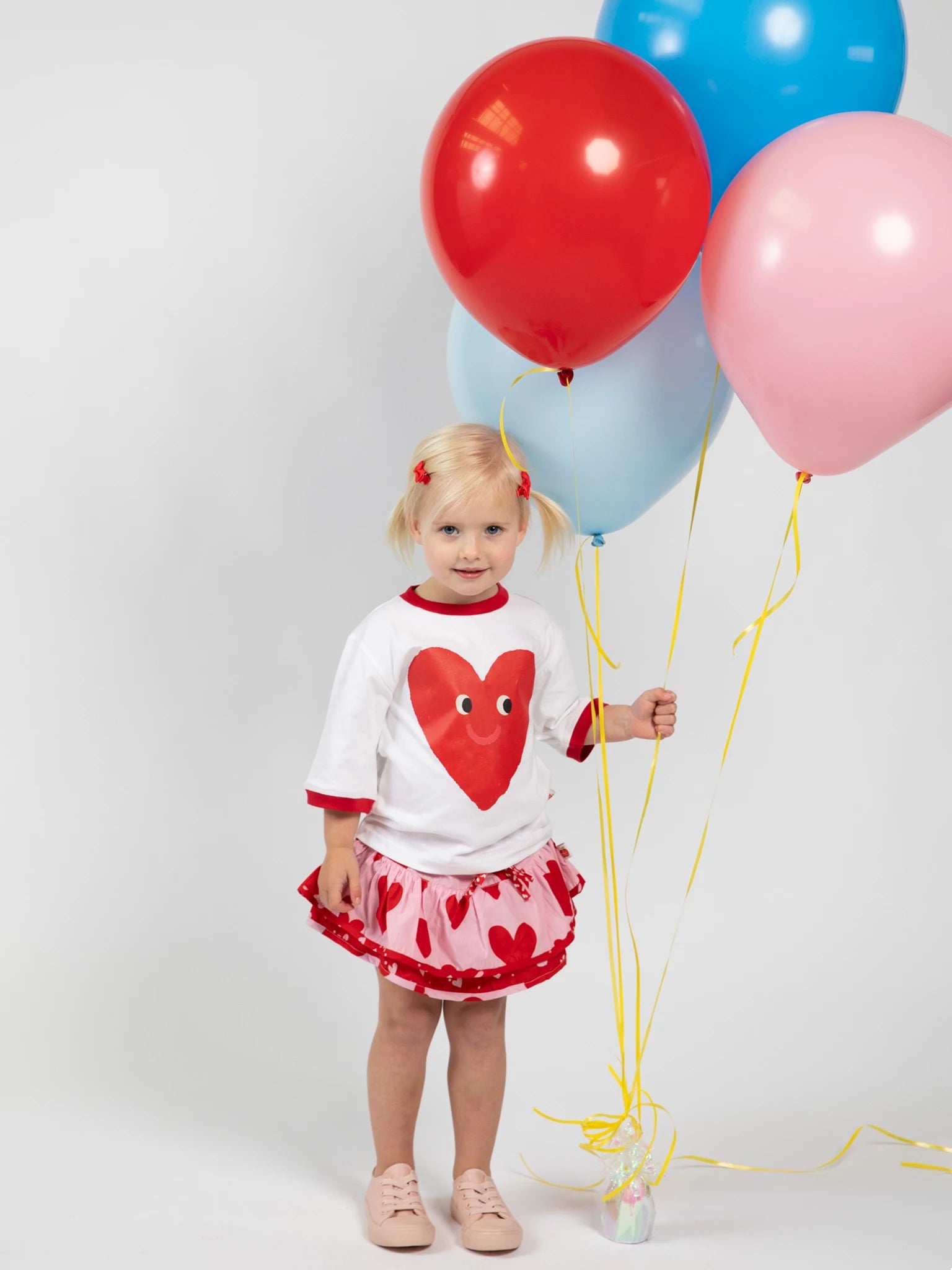 A young girl holds three balloons, wearing the Essentials Happy Heart Tee with Red Binding, an oversized heart-patterned skirt, and pink shoes, set against a plain light background.