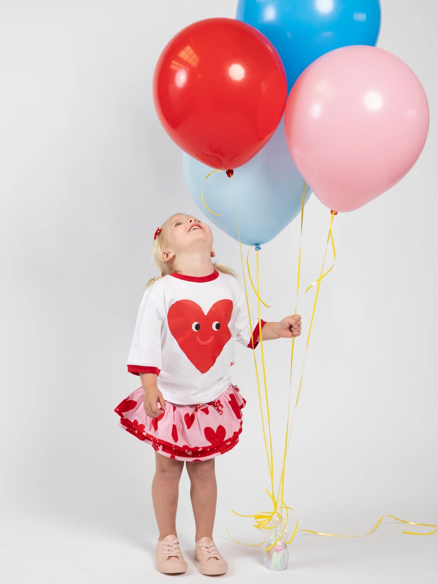 A young child wears the Essentials Happy Heart Tee with Red Binding, featuring an oversized fit, and holds red, pink, and blue balloons while gazing up at them against a plain white background.