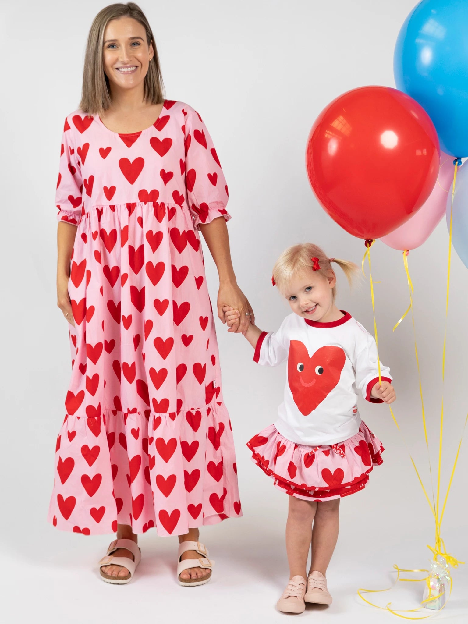 A woman and a young girl stand together; the woman wears an oversized fit pink dress with red hearts, while the girl wears the Essentials Happy Heart Tee with Red Binding and holds red and blue balloons.