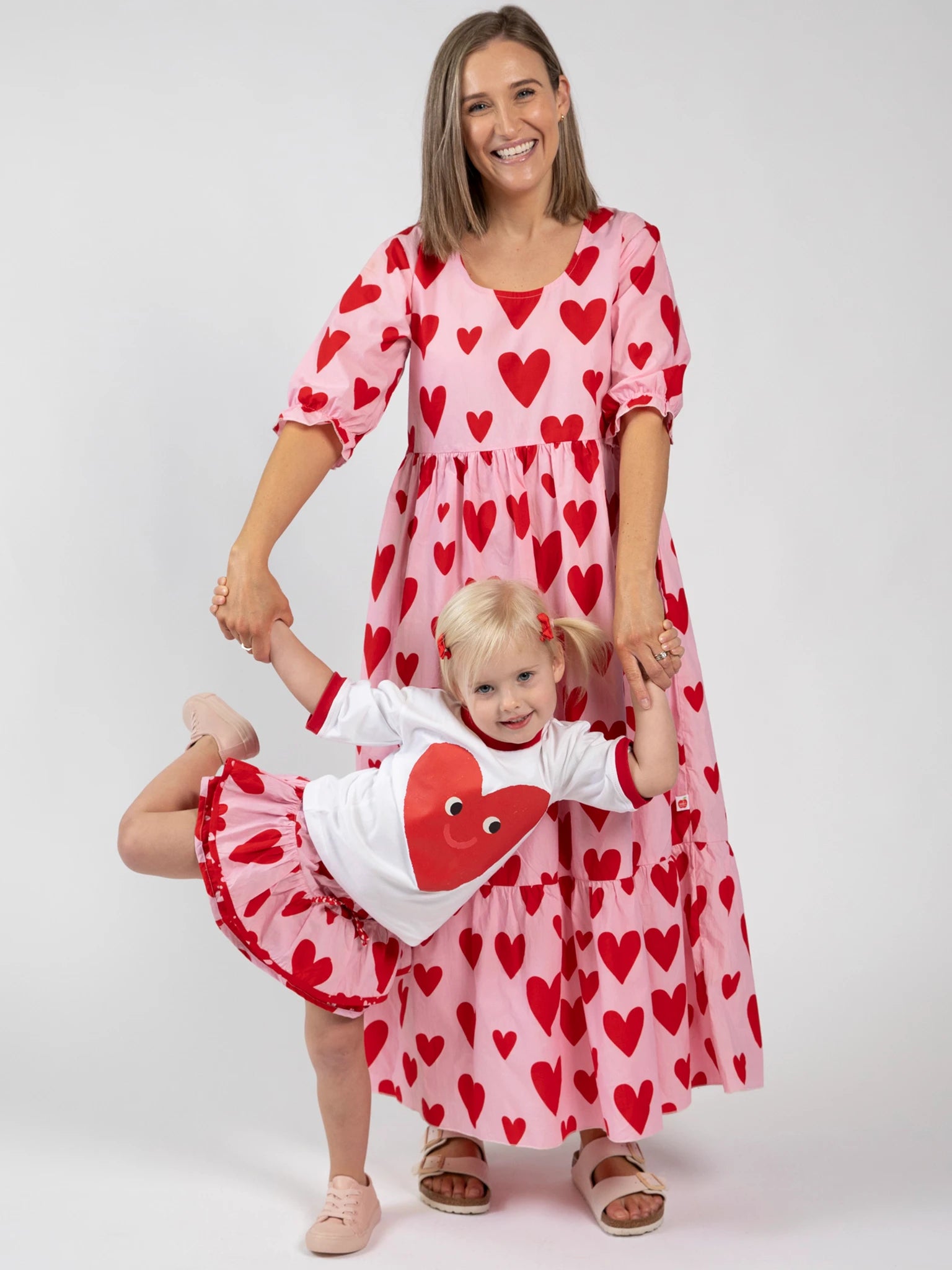 A woman and young girl wear matching hand-printed cotton outfits with red hearts by Big Hearts. The girl poses in the Big Heart Blossom Skirt with Bloomer, standing on one leg as she holds her mom’s hands.