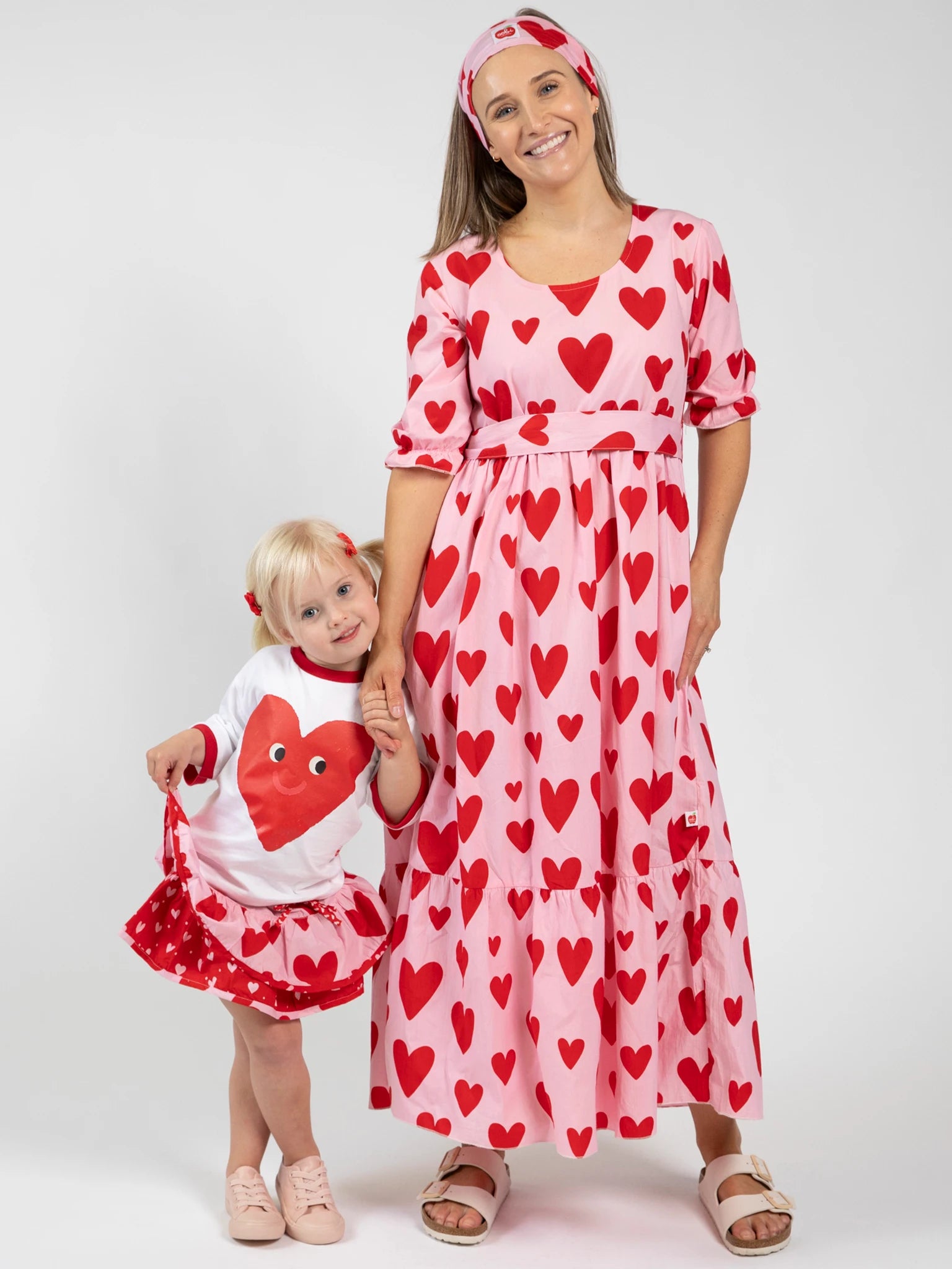 A woman and a child wear matching Big Hearts outfits, including the Women's Big Heart Maxi Dress, featuring red heart prints on white and pink backgrounds, as they pose against a plain white backdrop.