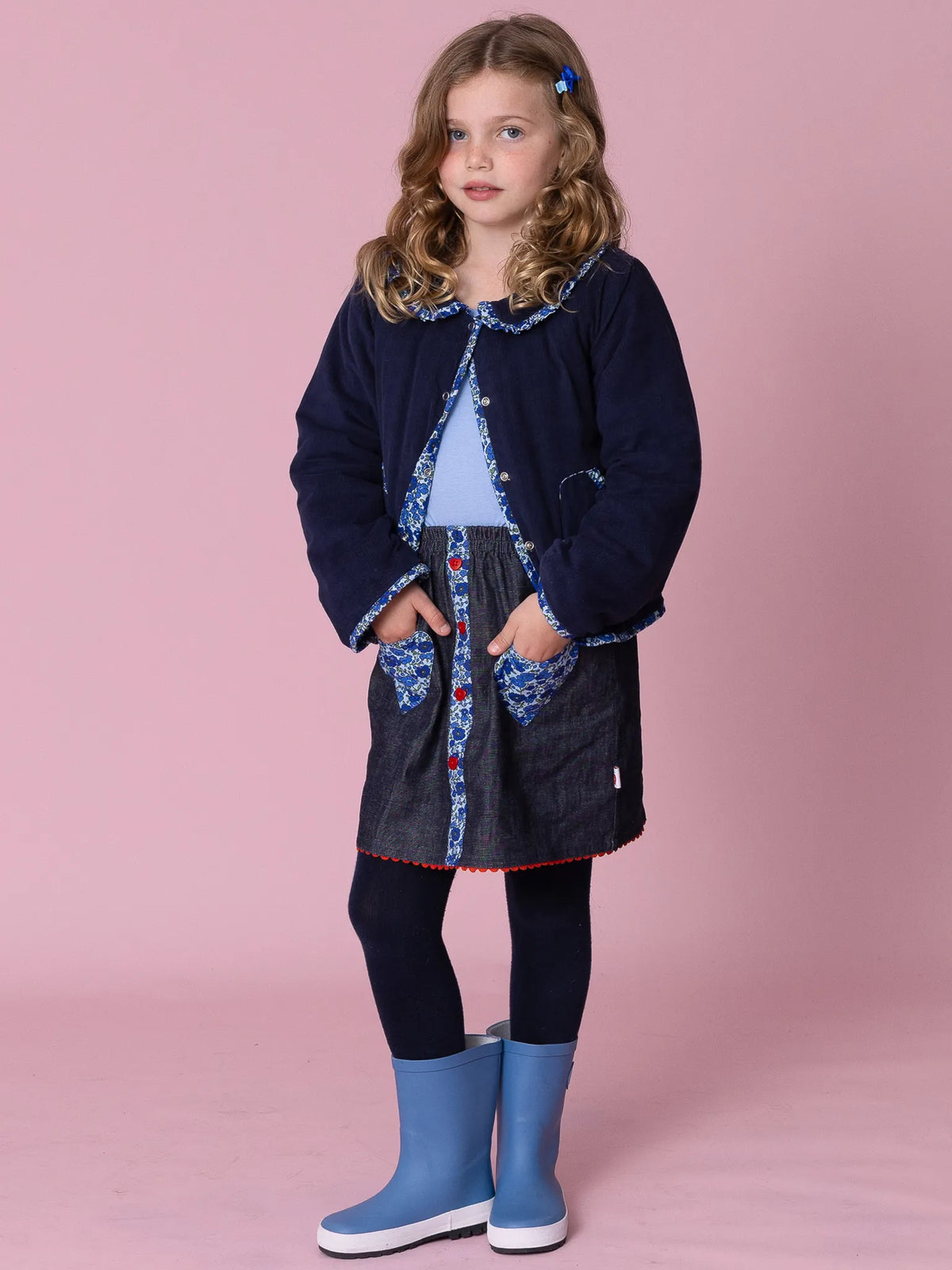 Young girl wearing a navy coat, denim skirt, and blue boots on a pink background