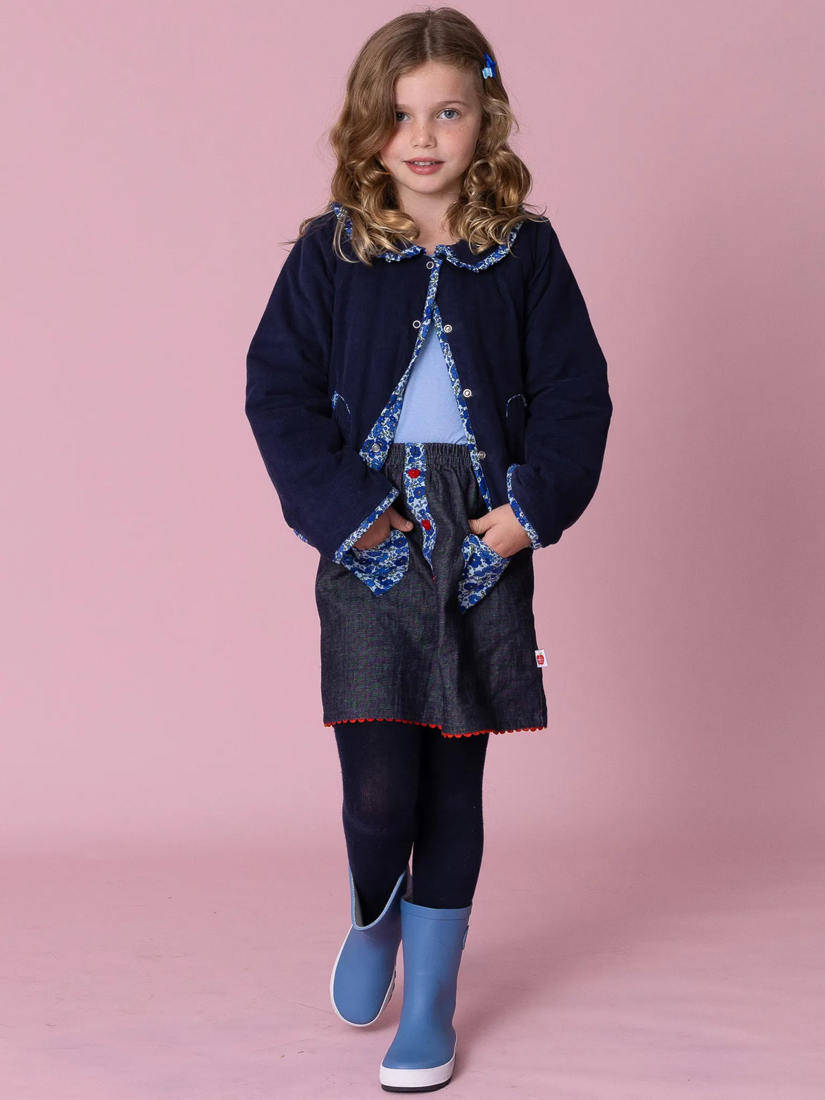 Young girl wearing a navy coat with blue accents, denim skirt, and blue boots on a pink background